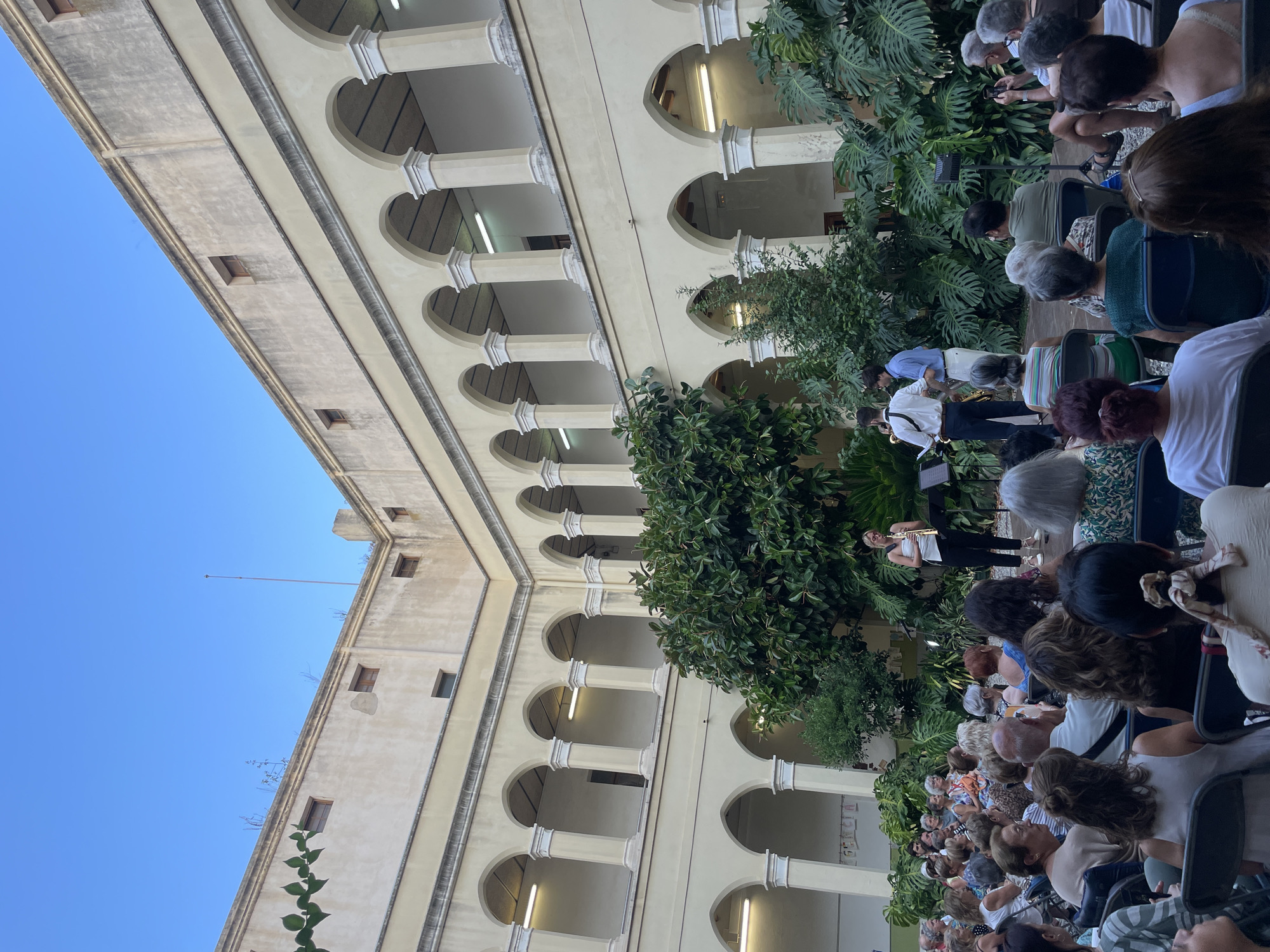 The image captures an outdoor performance in the cloister of the Sant Pere Church in Calella, Spain. A group of musicians, including a woman playing a clarinet and two men playing a saxophone, are performing on a small stage in the center of a courtyard. They are surrounded by lush green foliage. An audience of approximately fifty people, seated in chairs, is watching the performance. The cloister features classical architecture with arched walkways and columns. The sky above is a clear blue.