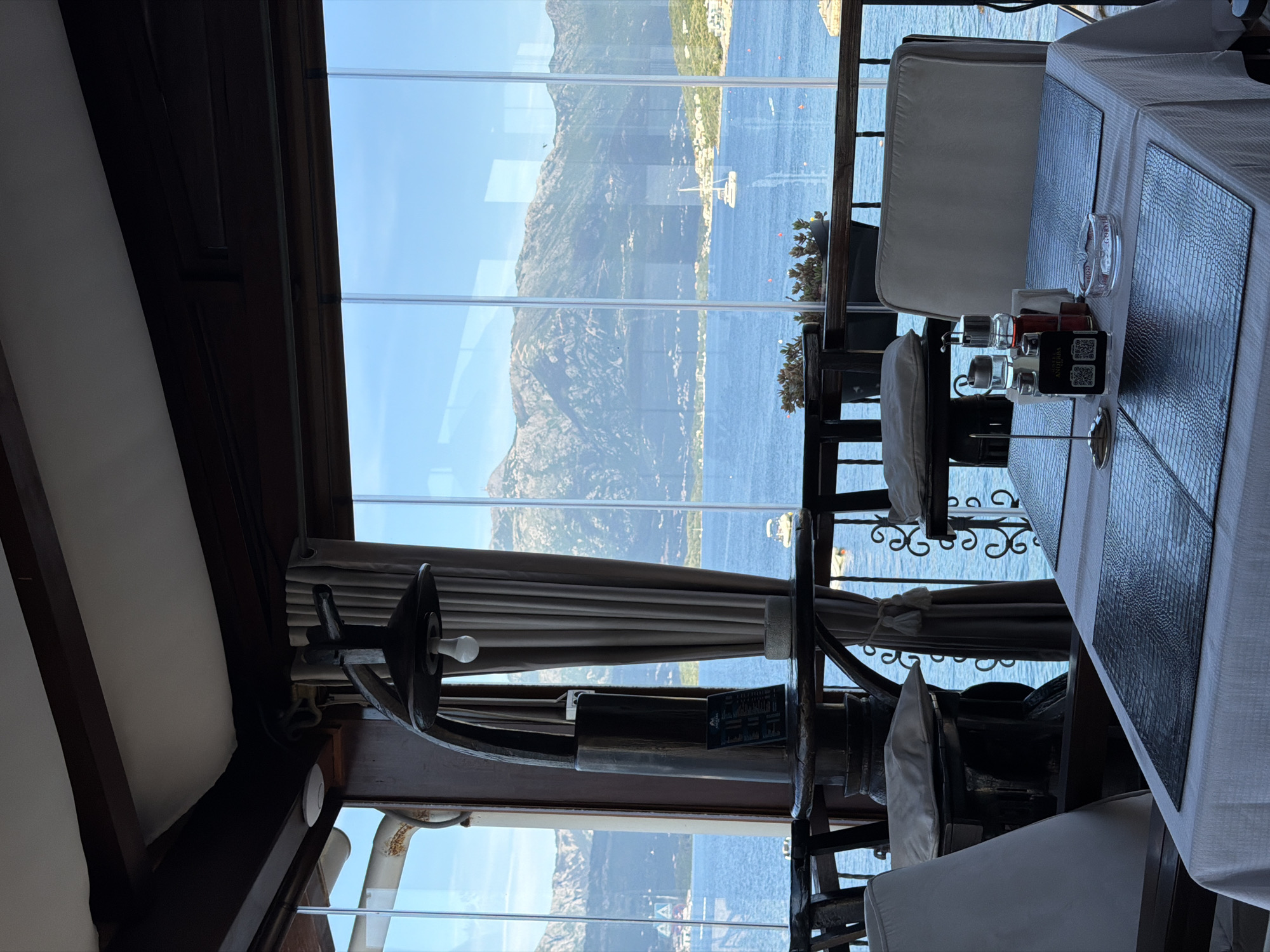 The image shows an interior view of a restaurant in Krašići, Montenegro, overlooking the Bay of Kotor. A large, vertically-oriented window offers a view of the mountains, the sea, and the town. Tables with white tablecloths and reptile print placemats are set for dining. There are chairs with white cushions. A decorative helm mounted above the window adds a nautical theme to the space. Table accessories such as condiment holders and QR code stands are present on the tables.