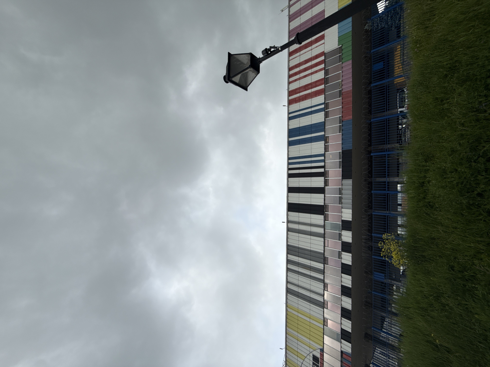 A tilted eye-level shot displays a street lamp mounted on a building with a colorful striped facade, located in Khimki, Russia. The sky is overcast with gray clouds. Lush green grass lines the right edge of the frame, obscuring part of a blue-railed structure attached to the building. The striped building facade is composed of horizontal bands of varying colors, including red, white, blue, gray, black, and yellow. The street lamp is black and traditional in design, extending from a pole attached to the building's side. The windows of the building can be seen in the stripes.