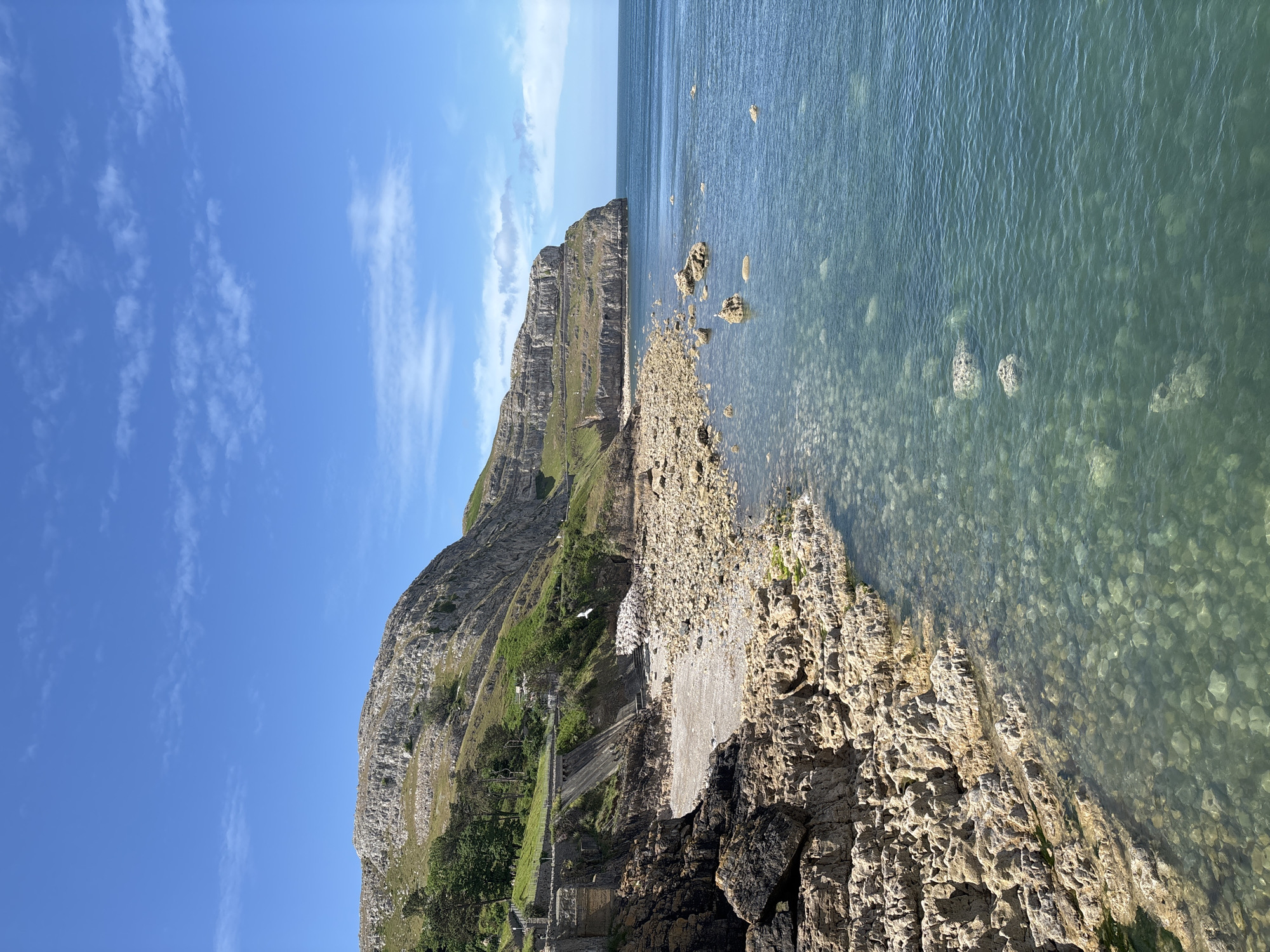 The image captures a coastal scene in Llandudno, United Kingdom. A rocky shoreline transitions into clear, shallow water with visible pebbles at the bottom. Adjacent to the water is a cliff face, with vegetation and a set of stone steps visible. The sky above is blue with wispy clouds.