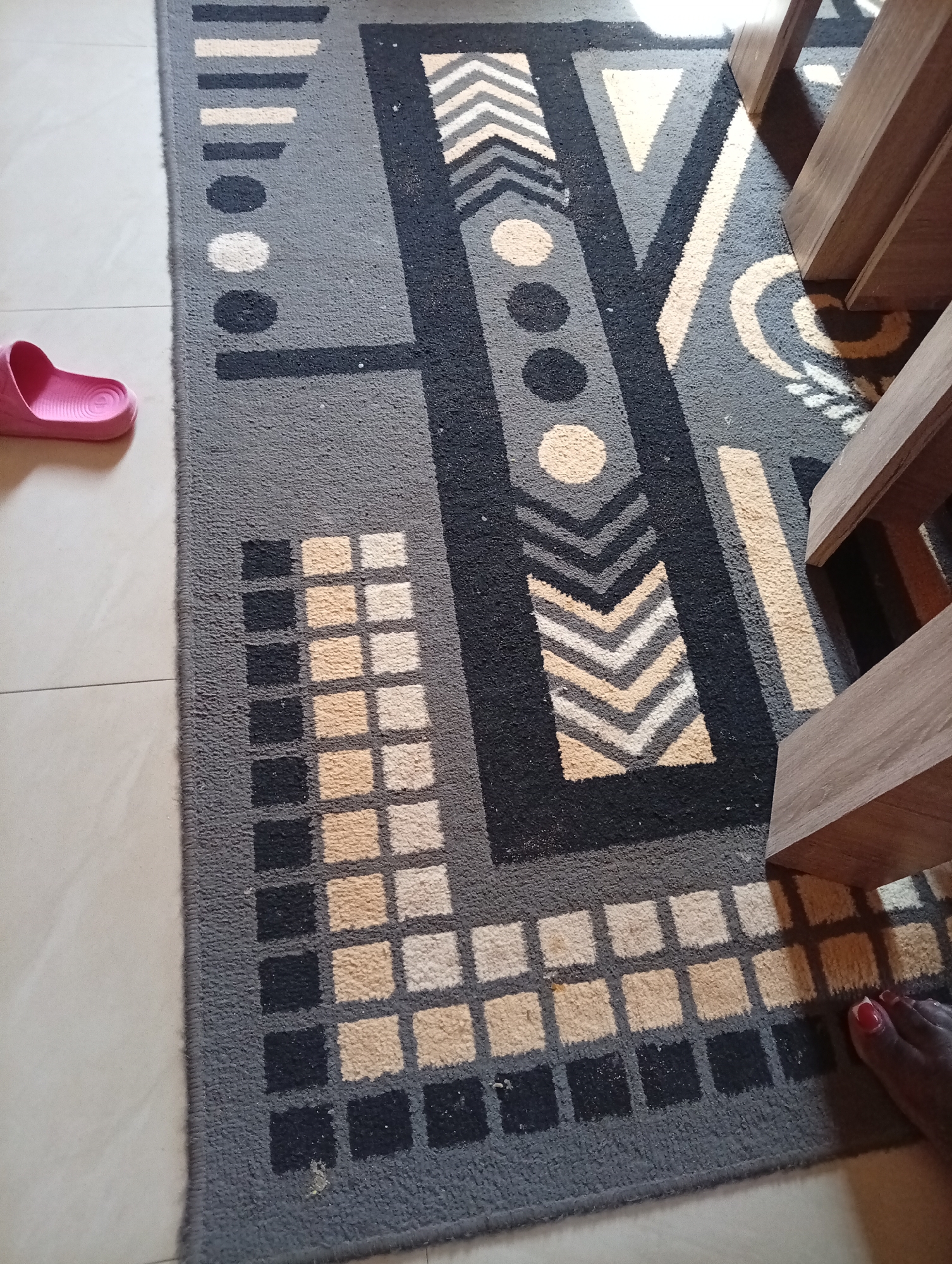The image shows a close-up of a gray rug with a geometric pattern. The rug features blocks of black, cream, and dark gray shapes, including stripes, circles, and chevrons. A pink flip-flop is visible on the tiled floor to the left of the rug. In the upper right corner, the legs of wooden furniture are visible, casting shadows on the rug. A person's foot with red nail polish is at the bottom right of the rug. The setting appears to be indoors. The location in Gura Top, Nigeria is not indicated by any visual cues in the image.