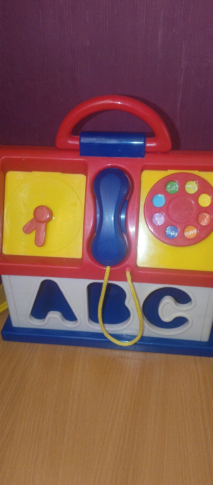 This is a close-up, eye-level shot of a colorful, plastic baby toy, likely an activity cube, set against a muted, ribbed purple background. The toy is predominantly red, with sections of bright yellow, blue, and white. The top of the toy features a red, curved handle and a blue plastic piece that appears to be part of a phone receiver.
The front of the toy has three distinct sections. The leftmost section is a yellow square with a red, clock-like dial featuring a single red hand. The middle section is a blue, elongated oval shape, potentially a button or a speaker. The rightmost section is another yellow square containing a red, circular dial with eight colored buttons arranged around its perimeter, resembling a telephone keypad with buttons in red, orange, green, light blue, and purple.
Below these top sections, the front of the toy displays three large, cut-out shapes in white, spelling out "A," "B," and "C" from left to right. These are likely openings for shape-sorting activities. A yellow rope is attached to the toy and hangs down, forming a loop. The toy rests on a light brown, wooden surface that appears to be a tabletop or floor. The lighting is even, and there are no obvious shadows, suggesting an indoor setting. The overall impression is one of a well-used, playful toy.