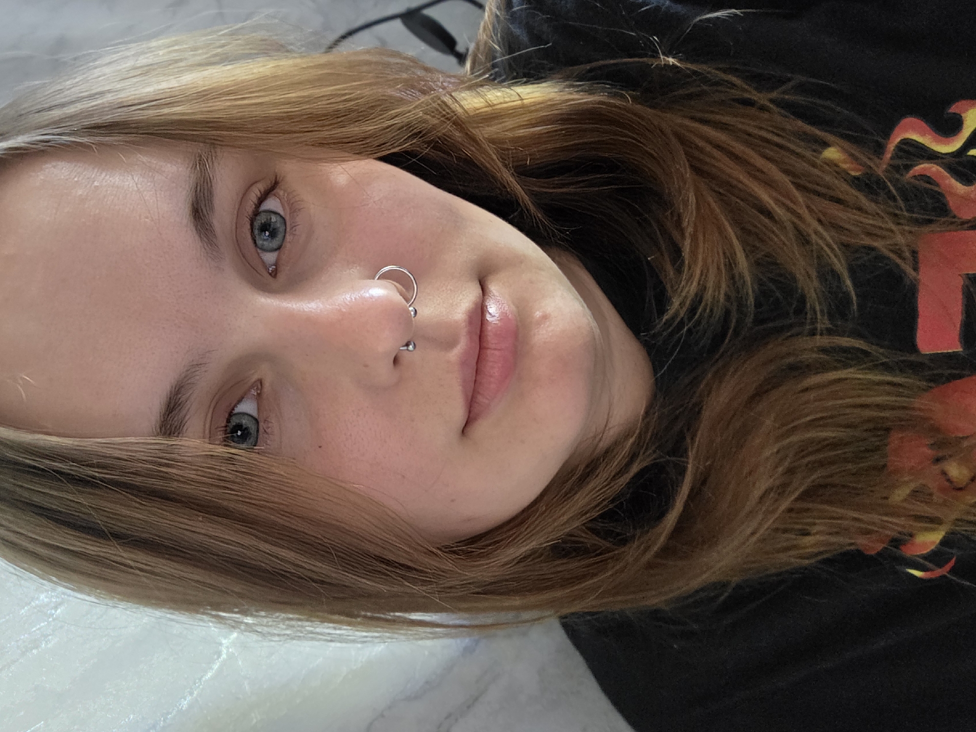 A close-up, selfie-style photograph shows a person with light brown hair and light blue-grey eyes. The person is facing the camera with a slightly tilted head. A silver nose ring is visible in the left nostril, and a smaller silver stud is in the center of the nose. The person is wearing a black shirt with a design that includes red and yellow flames. The background is a blurred white surface. The location is Unknown, Unknown.
