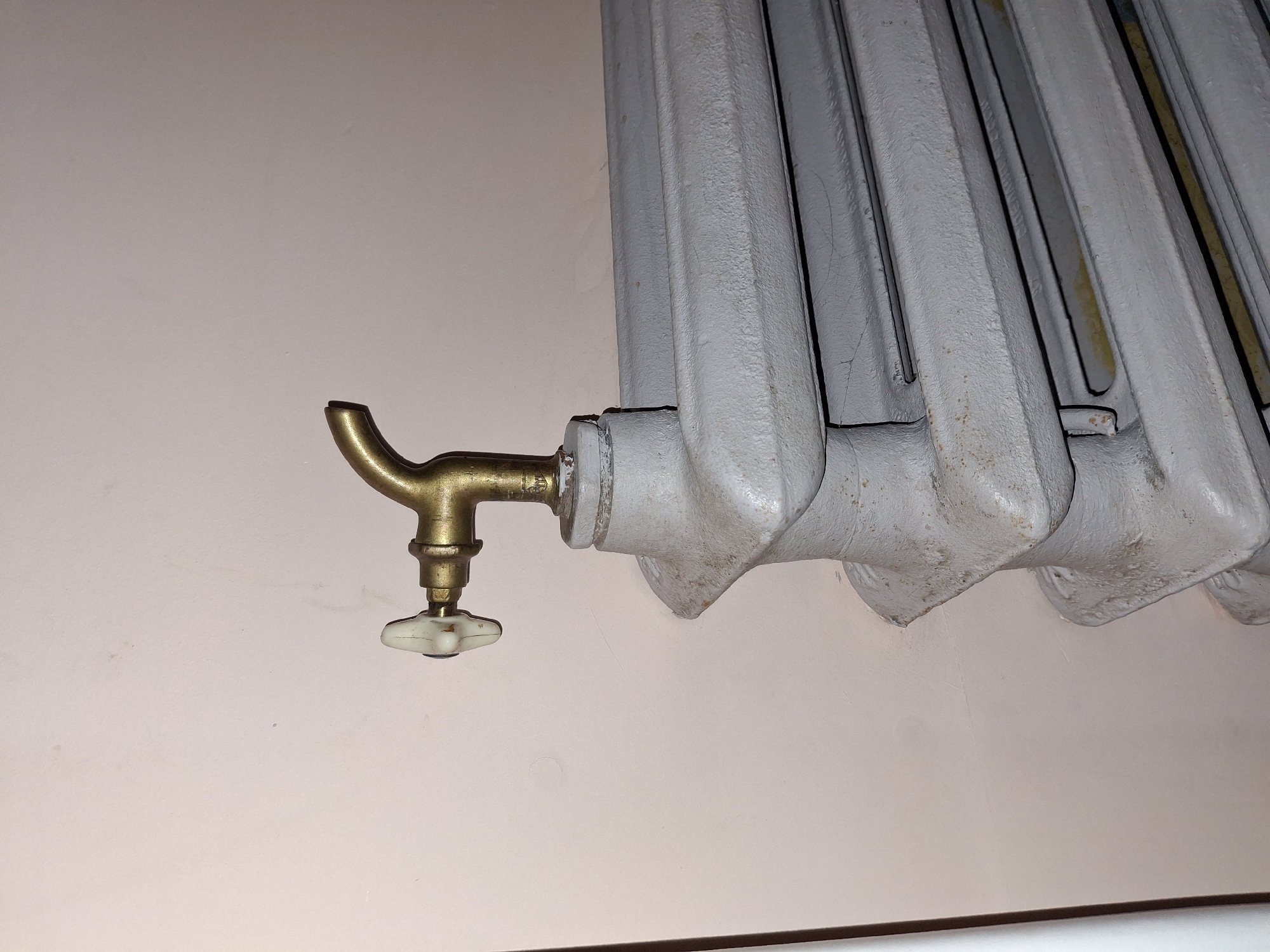 This media file is a close-up photograph of a radiator with an attached brass valve. The radiator is painted light gray and appears to be made of cast iron, with multiple vertical sections. The valve has a curved brass spout and a white ceramic handle. The background is a plain, light-colored wall, possibly painted. There are no people or other objects clearly visible. The lighting suggests an indoor setting, possibly a room in a home or building. The image focuses on the functional components of the heating system. No text is visible in the image. The location context provided is Ogre, Latvia.