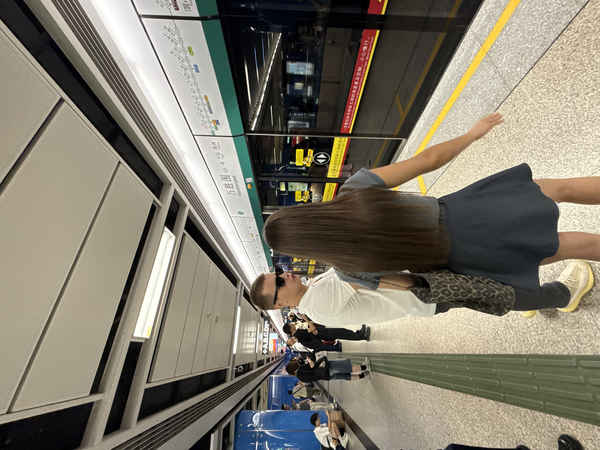 The image captures a scene at a subway station in Haizhu District, China. In the foreground, a woman with long brown hair, wearing a dark blue dress, stands facing away from the camera, her right arm extended slightly. Beside her, a man with short hair and sunglasses, dressed in a white polo shirt and dark pants, looks towards the woman. He carries a leopard-print bag on his left shoulder. They are positioned on a light-colored, speckled platform, adjacent to a train with closed doors. A bright yellow line marks the edge of the platform. Behind the train doors, the interior is visible, with reflections of the station's lights and signage. Overhead, a white ceiling with recessed panels and fluorescent lighting is prominent. The station platform features tactile paving strips in a dark green color. In the background, other passengers are visible, some standing and others seated or crouching, interacting with their phones or resting. Signs with Chinese characters and directional arrows are displayed on the wall above the train doors. The overall environment suggests a busy urban transit hub.