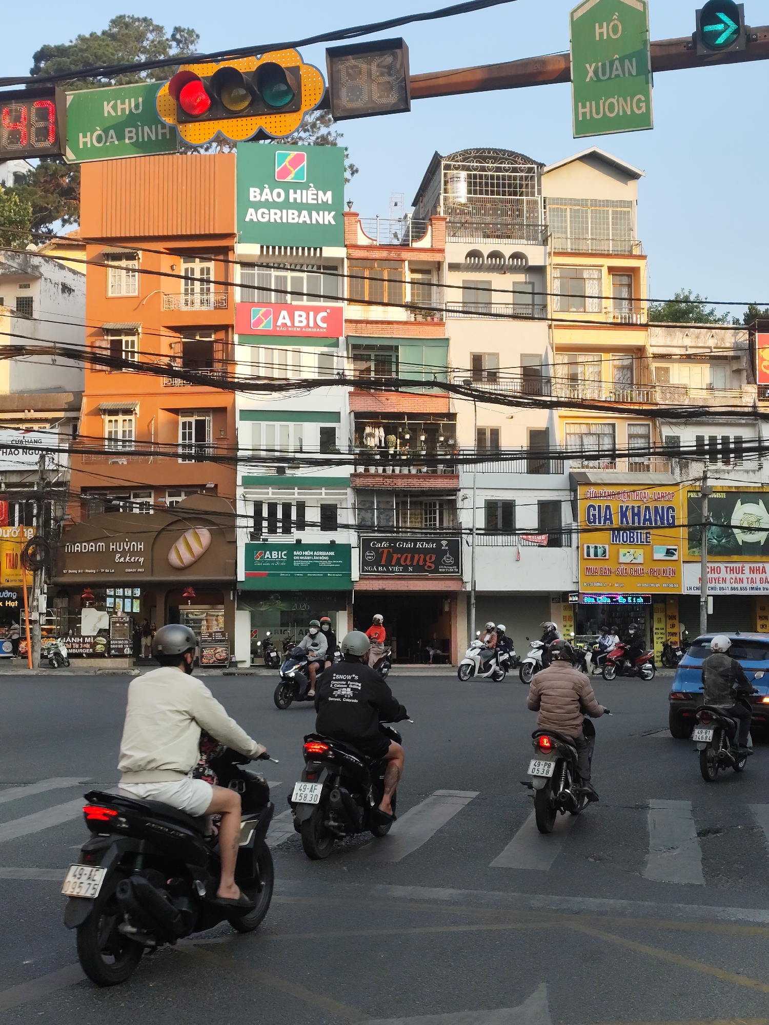 The image shows a busy intersection in Da Lat, Vietnam. The intersection is marked by zebra stripe crosswalks. Several motorbikes are in the intersection, either moving or stopped. There is also a blue car in the intersection. Buildings with a variety of commercial signs line the streets around the intersection. The signs include: "KHU HOA BINH", "HO XUAN HUONG", "BAO HIEM AGRIBANK", "ABIC", "MADAM HUYNH Bakery", and "GIA KHANG MOBILE." Above the intersection, a traffic light shows a green arrow for turning. People on motorbikes are wearing helmets. There are electrical wires crisscrossing the scene. The sky is clear.