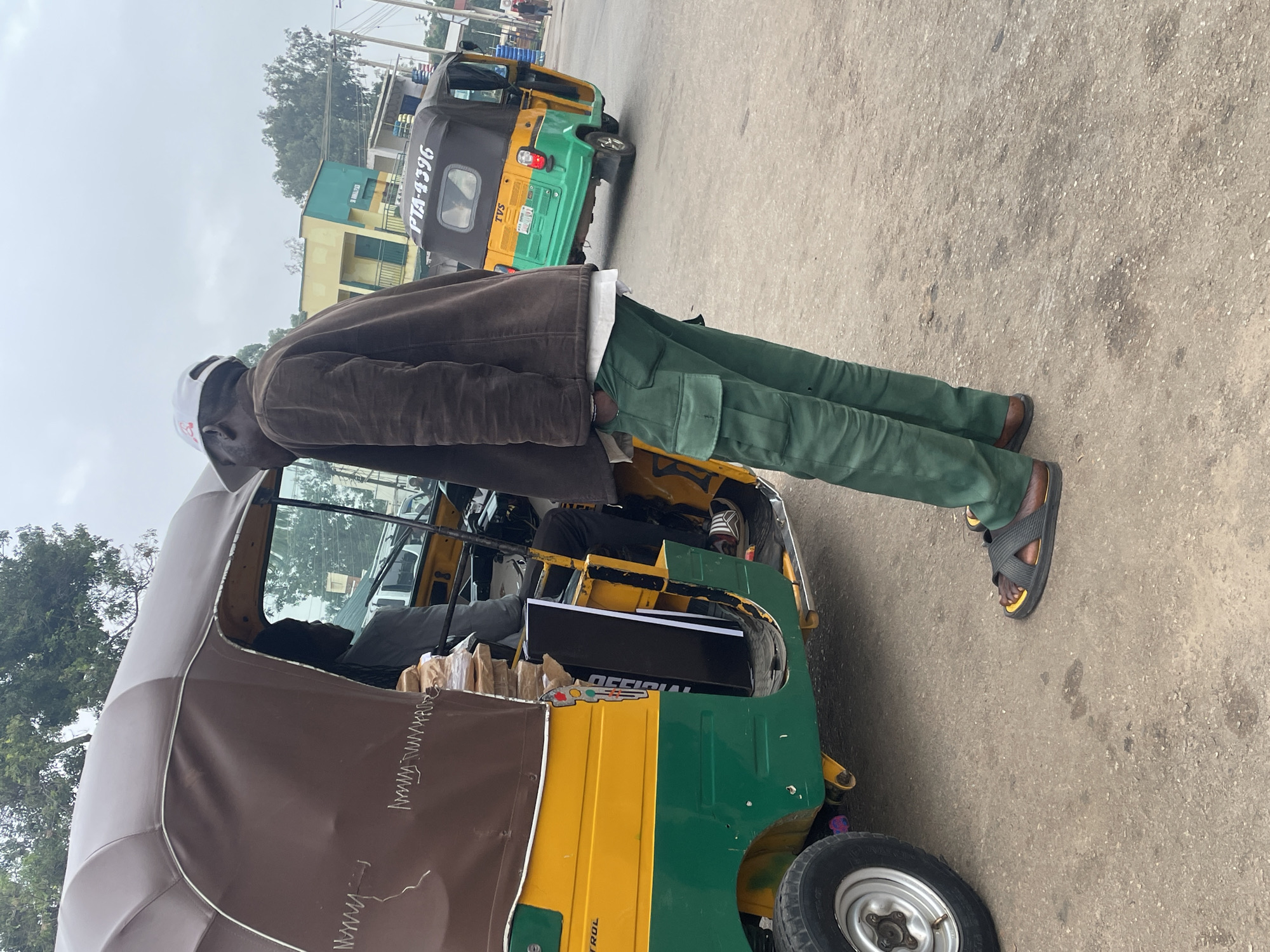 In Jos, Nigeria, on a cloudy day, a man wearing a brown jacket, green cargo pants, and black and yellow sandals stands next to a yellow and green auto-rickshaw. The auto-rickshaw, with a brown canopy, is filled with stacks of what appear to be books or documents. Another person is visible inside the auto-rickshaw. In the background, another auto-rickshaw with license plate PTA-4396 is parked, and buildings with colorful facades line the street. The ground is paved asphalt.