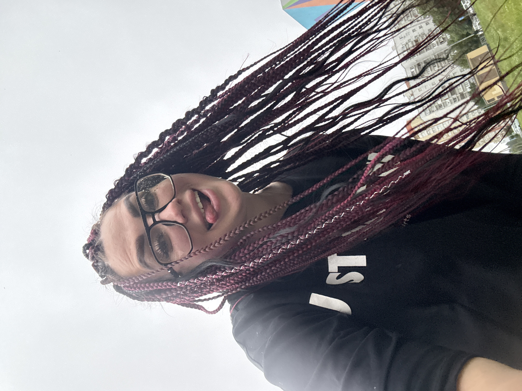 A young woman with dark braided hair, some of which is a deep maroon color, is captured in a close-up, slightly upward-angled selfie. She is wearing black-rimmed glasses and a black sweatshirt with white lettering on the chest that appears to say "JUST." Her tongue is sticking out, and her mouth is open in a playful expression. The background is overcast and indistinct, but hints of a residential area with apartment buildings and greenery are visible. The scene is likely outdoors, as suggested by the natural lighting and the faint presence of buildings and a grassy area, possibly in a park or a courtyard in the city of Tolyatti, Russia.
