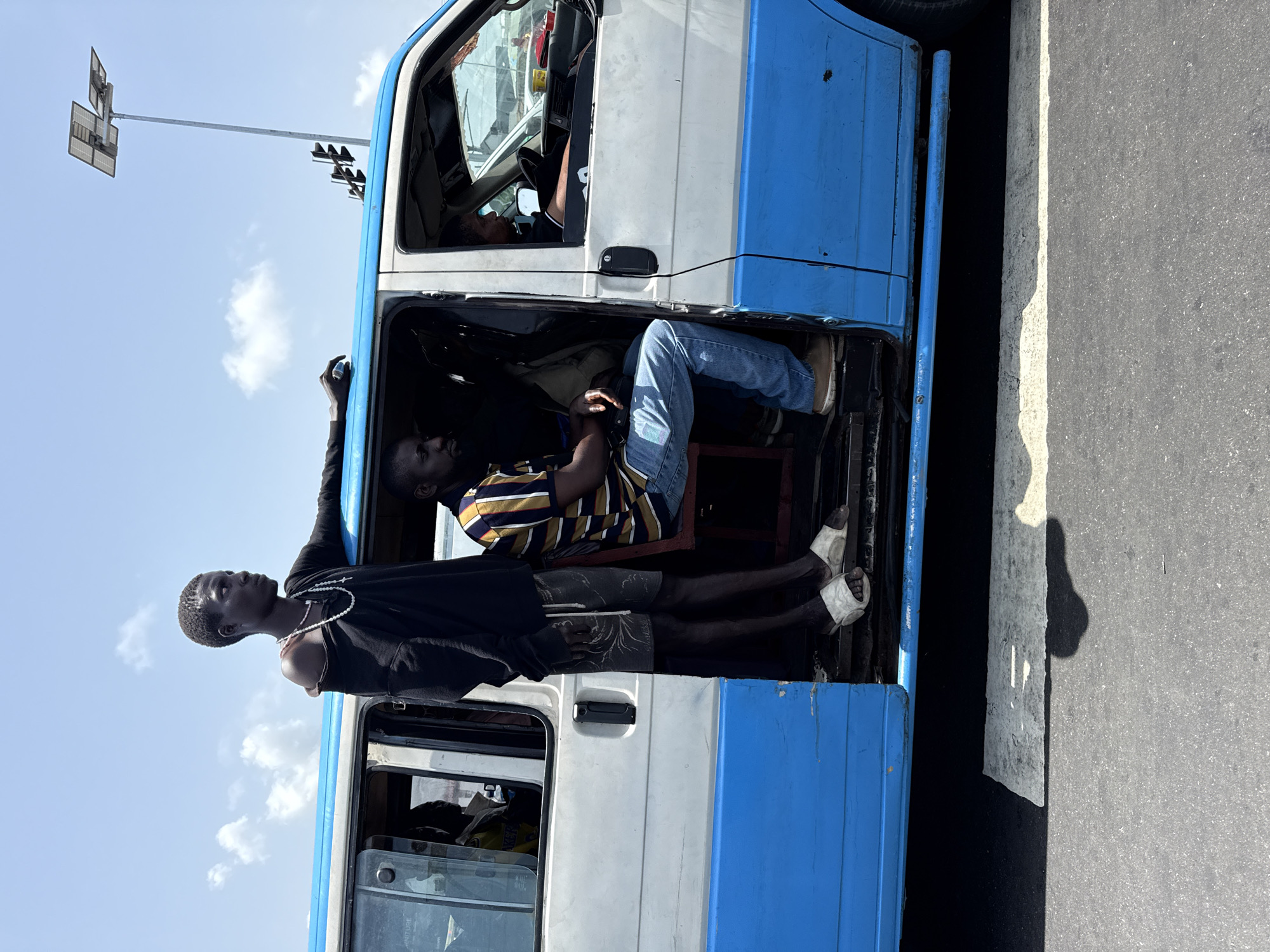 A medium, eye-level outdoor shot depicts a blue-and-white minibus in Lekki, Nigeria. Three individuals are visible. One person is driving, one seated inside, and another stands in the doorway. The standing individual, wearing a black long-sleeved shirt, shorts, a white necklace, and sandals, holds the bus. The seated person is wearing a striped shirt, jeans, and brown shoes. Part of the asphalt road is visible, along with a tall, metal pole with what appears to be solar panels or a similar structure attached. The sky is pale blue with scattered clouds.