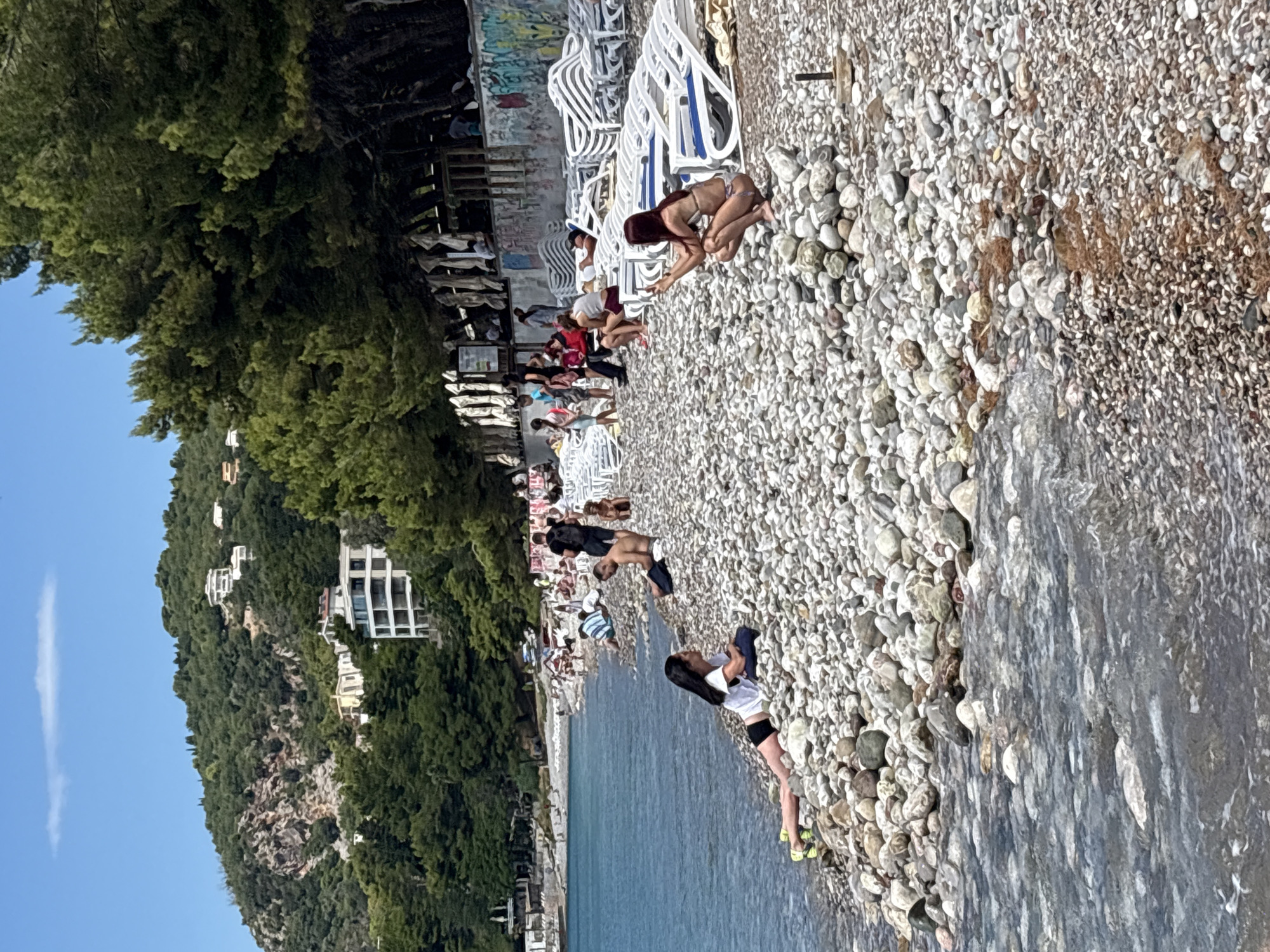 The image depicts a crowded pebble beach in Bar, Montenegro. The foreground shows the clear, shallow waters of the Adriatic Sea lapping against the shore, with a woman in a white t-shirt and black shorts lying on her side on the pebbles, her left leg extended towards the water. Further up the beach, a woman in a bikini is crouched down, holding up her phone as if taking a picture. In the background, numerous people are scattered across the beach, some sitting on towels, others on lounge chairs. A line of white lounge chairs stretches along the upper portion of the beach, many of them unoccupied. Behind the chairs, a wall with graffiti is visible, and beyond that, dense green trees line the shore, with buildings and houses nestled into the hillside of the mountainous terrain. The sky is a clear blue with a single, wispy cloud. The overall scene conveys a vibrant summer day at a popular tourist destination.