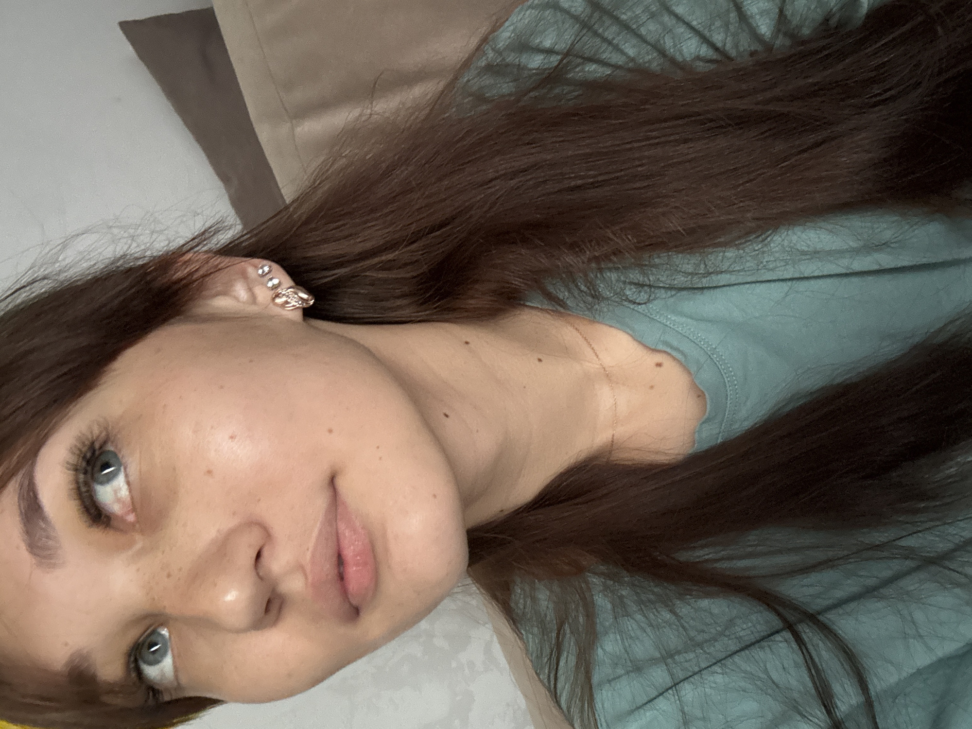A close-up, medium shot captures a young woman from the chest up, positioned slightly to the left of the frame. She has fair skin, visible freckles across her nose and cheeks, and bright blue eyes looking upwards and to the left. Her lips are slightly parted and appear soft and pink. She has long, dark brown hair that drapes over her right shoulder and down her chest. She is wearing a teal t-shirt and several earrings in her left ear: a small stud and a larger, more elaborate earring with a bow-like design and small stones. A thin gold necklace is visible around her neck. The background is blurred, showing a textured light gray wall on the left and a brown upholstered surface to the right, possibly a sofa or chair, with a shadow forming a diagonal line. The lighting appears soft and natural. The image is a selfie taken from a slightly elevated angle.