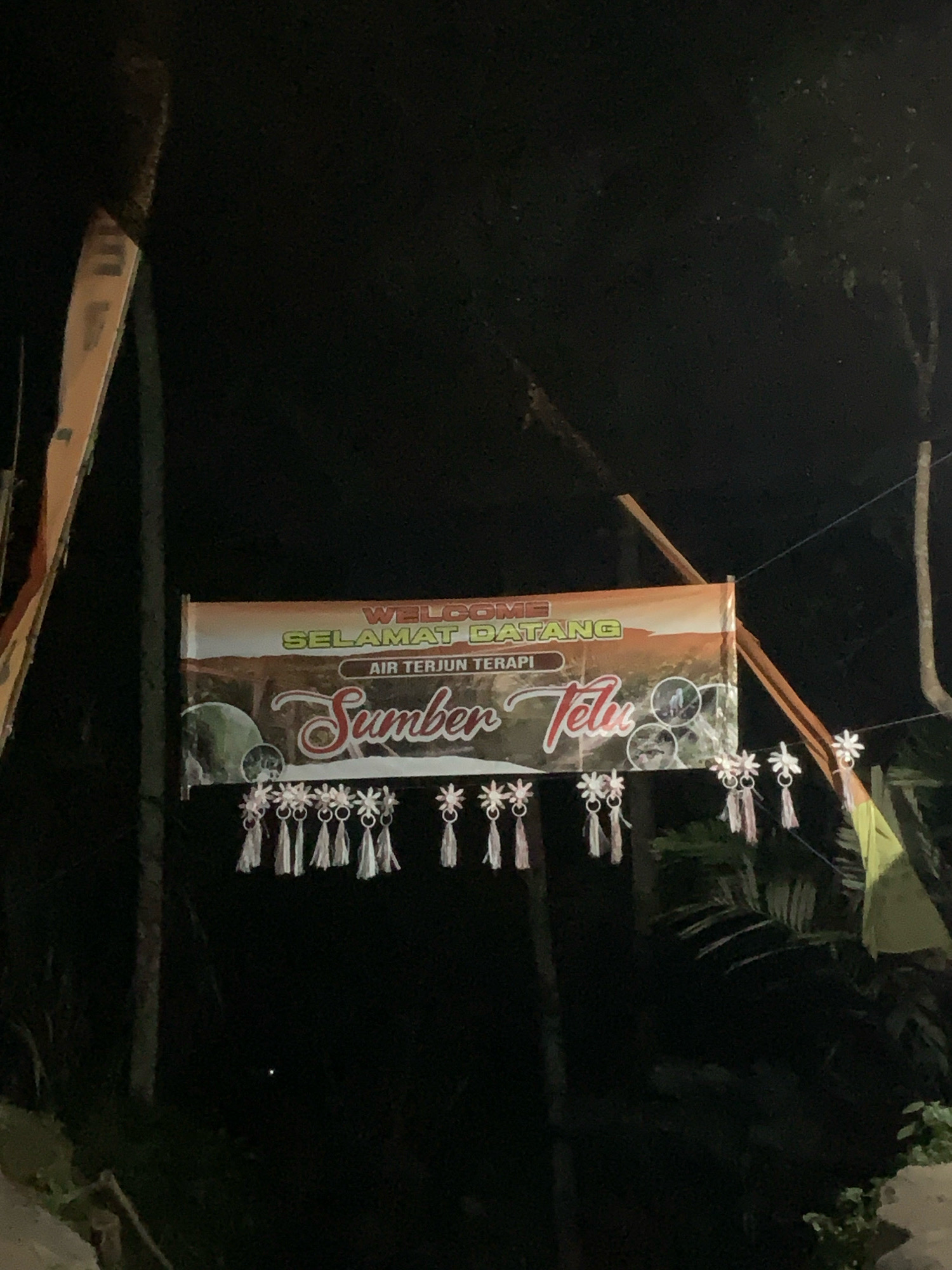 An illuminated banner hangs between trees at night, likely in Pronojiwo, Indonesia, advertising "Sumber Telu" and "Air Terjun Terapi". The banner, featuring an image of a natural landscape with a waterfall, is adorned with decorative tassels and star-shaped ornaments. The background is dark, suggesting it is nighttime, with some foliage and tree trunks visible.