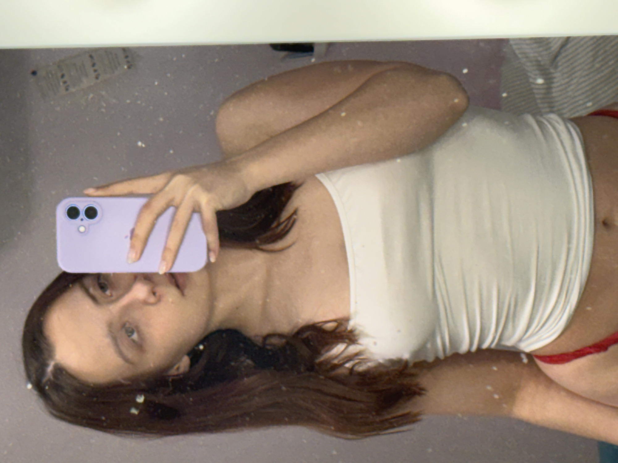 A young woman is taking a selfie in a mirror. She is holding a purple iPhone in her left hand, partially obscuring her face. She has brown hair and is wearing a white tank top and red underwear. The mirror is speckled and reflects a glimpse of striped bedding and various items. The location is unclear, and there is no verifiable connection to Прикамский, Russia.
