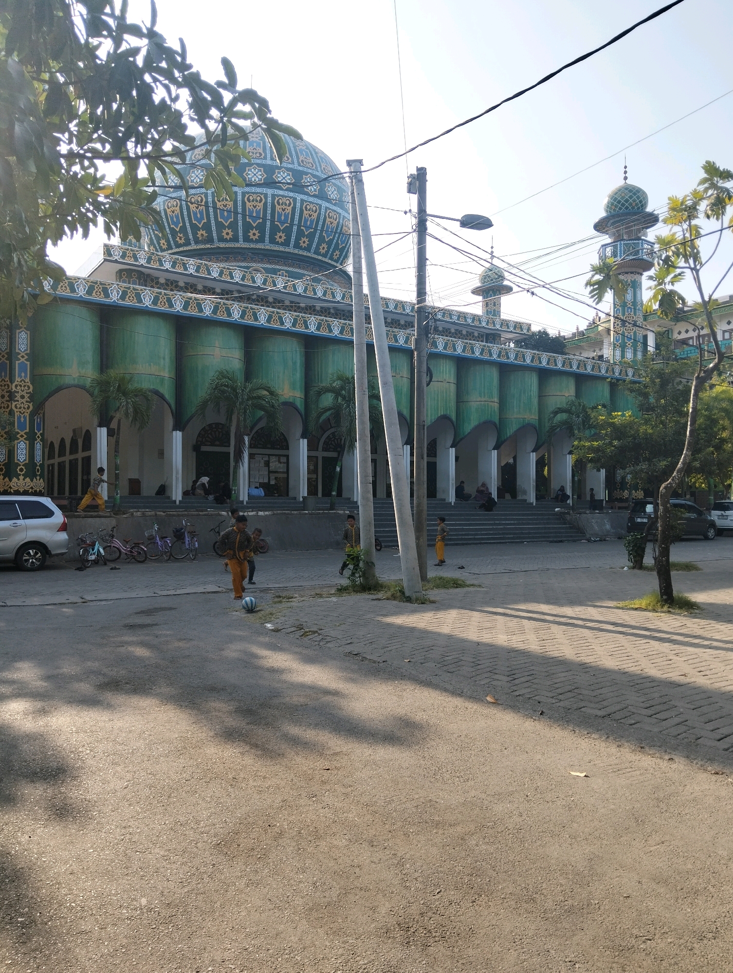 The image shows a mosque in Paciran, Indonesia, under a clear sky. The mosque has a large, teal-colored dome with intricate golden and blue patterns. The building features a green facade with arched entrances supported by columns. Several palm trees are planted in front of the mosque. In the foreground, there is a paved area with several people, including children playing with a ball. Bicycles are parked to the left, and vehicles are visible on the right side of the street. Electrical poles and wires are present in the scene. The lighting suggests it is daytime, with shadows cast by trees and structures.