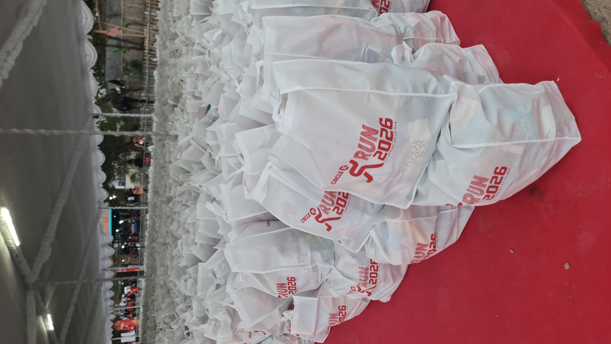 The image depicts a large collection of white fabric bags with red lettering, arranged in stacks on a red carpeted surface, likely for distribution at an event. The bags feature a logo with "CIRCLE K RUN 2026" and "SERIES OF CK RUN." In the background, beneath a white tent structure, a crowd of people is visible, suggesting an outdoor event or a preparation area for one. The environment appears to be an urban setting in Denpasar, Indonesia, indicated by the presence of people in casual attire and the general atmosphere of an event setup. The lighting is diffused, suggesting it's either daytime with overcast conditions or under artificial lighting within the tent. The focus is primarily on the bags, with the background slightly blurred.