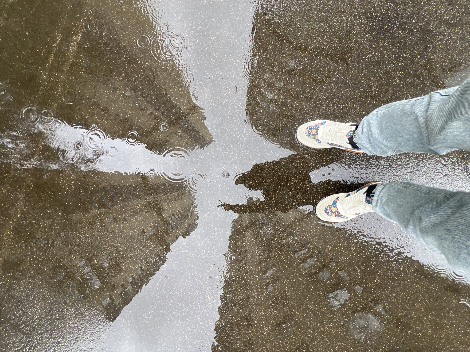 An eye-level shot shows a person standing in a puddle on a paved surface in Moscow, Russia. The puddle reflects buildings, creating a mirrored effect. Raindrops cause ripples in the water. The person wears light-wash jeans and white shoes with a geometric design. The pavement is wet and speckled.