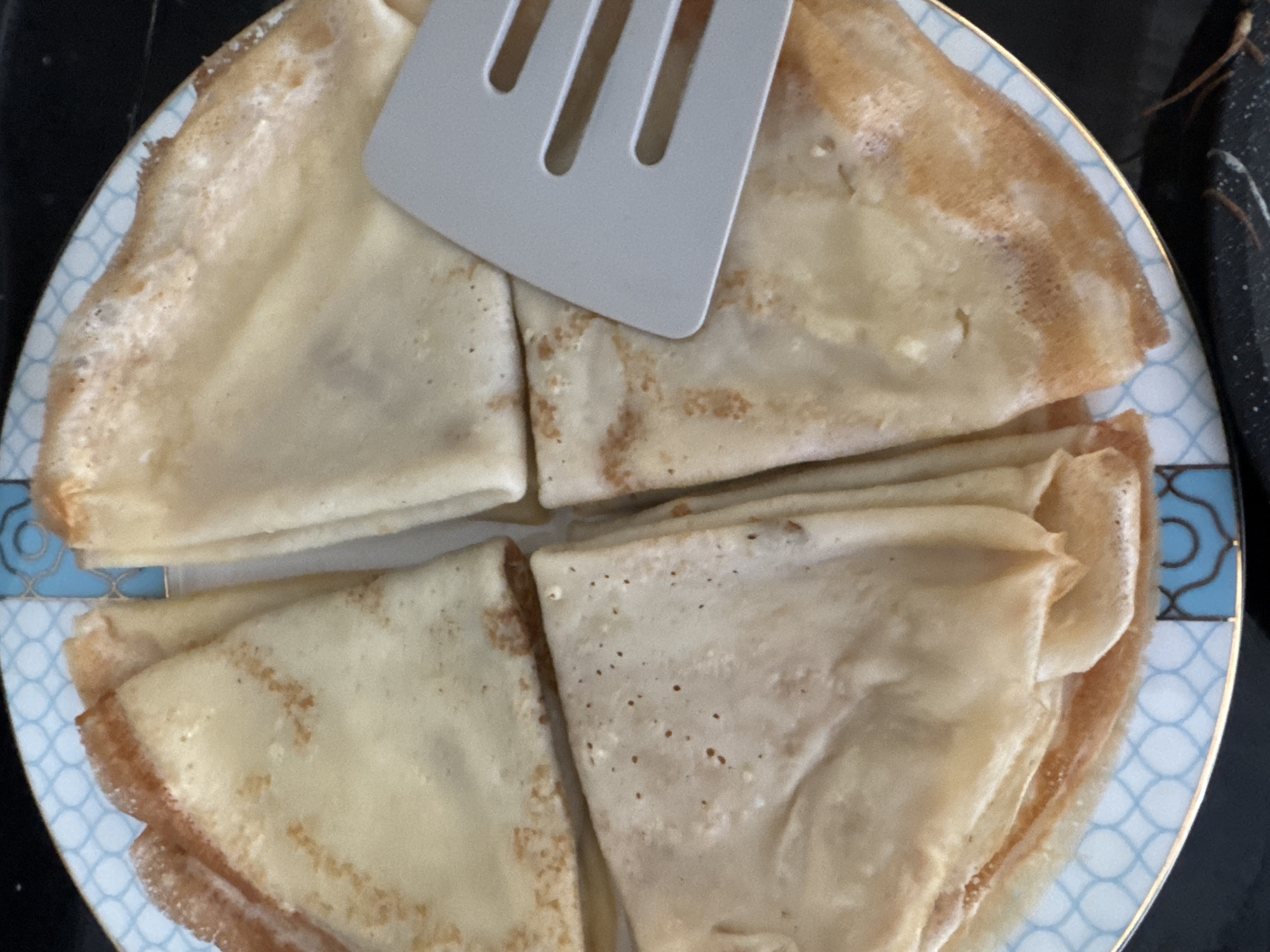 A top-down close-up view shows four folded crepes on a plate with a blue and white pattern. A light gray spatula with three horizontal slots is placed on top of the crepes. The crepes are golden brown with some darker, crispier edges. Some crepes appear slightly filled, showing a darker color inside. The plate has a thin gold rim. The background is dark and out of focus. The scene is set indoors, likely in a kitchen or dining area. There is no information to indicate the location within the city of Uyutnoye, Ukraine.