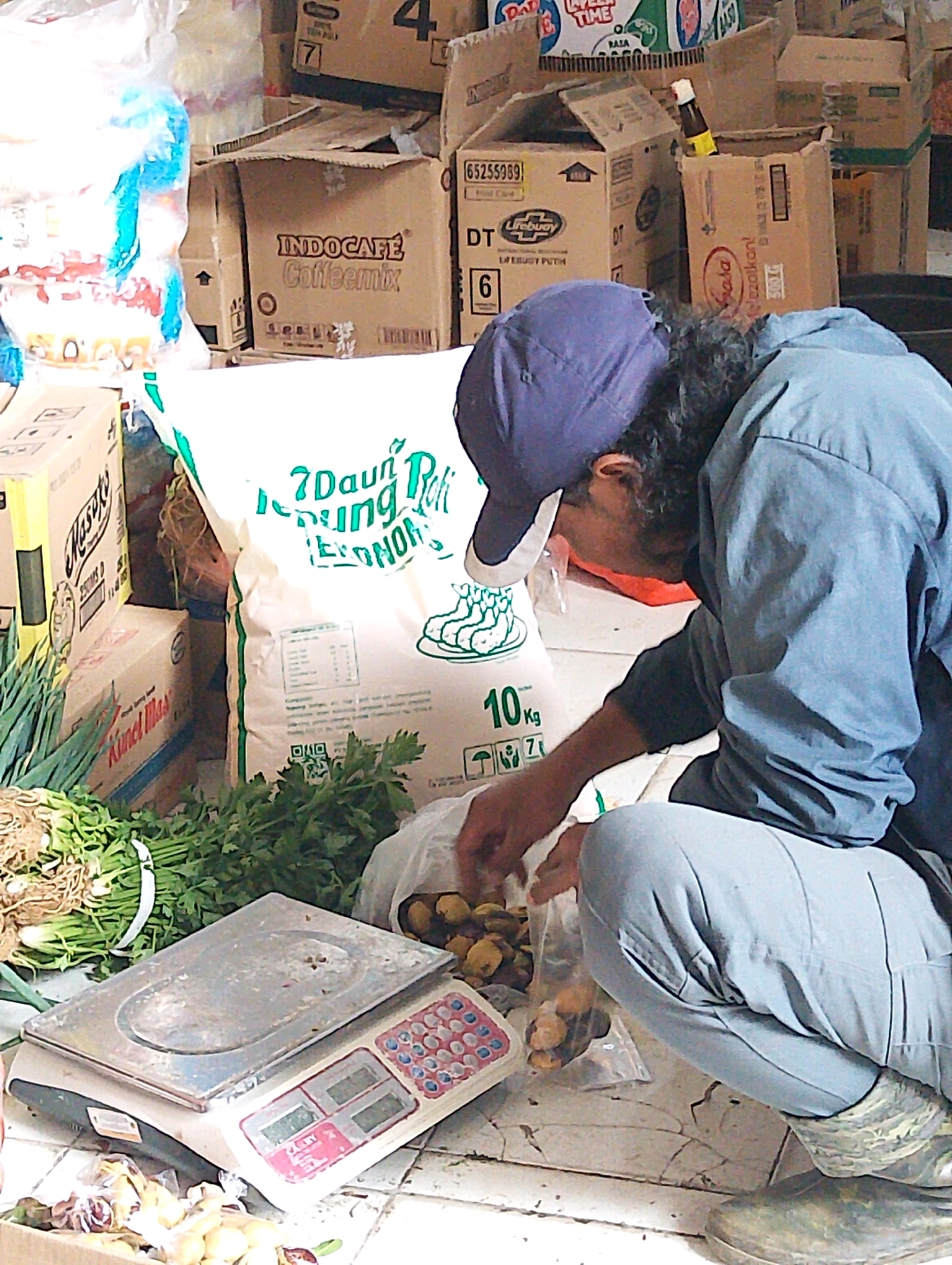 In Lembang, Indonesia, a man wearing a blue cap and a blue jacket is crouching down. He is in the process of sorting or weighing produce, possibly some kind of dried goods or nuts, from a clear plastic bag. To his left, a digital scale with a silver platform and pink buttons is visible. Beside the scale, a pile of green leafy vegetables and what appear to be onions or leeks are arranged. In the background, a large white bag with green text, likely containing rice or flour, is propped up. Behind the bag, several cardboard boxes are stacked, some bearing product labels such as "INDOCAFE Coffeemix" and "Masako". The environment appears to be an indoor market or a storage area, with a tiled floor and visible cracks.