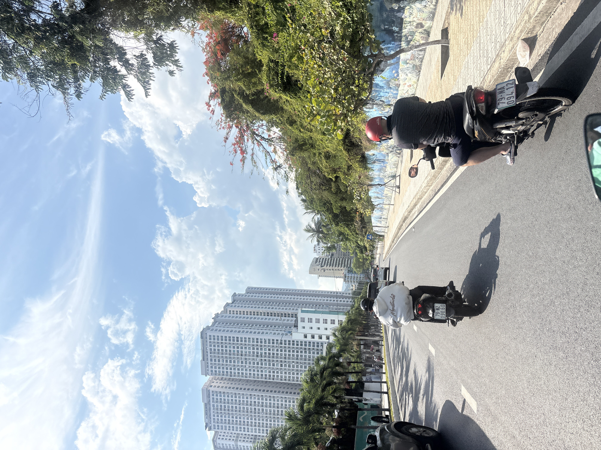 The image captures a street scene in Nha Trang, Vietnam, on a bright, sunny day with a partly cloudy sky. Two individuals are riding motorcycles in the foreground, moving away from the viewer. The rider in the foreground left wears a white hoodie and dark pants, with a white motorcycle and a license plate reading "79-N2 190 60". The rider in the foreground right wears a striped shirt, dark shorts, and a red helmet, on a black motorcycle with a license plate displaying "79-NA 094.12".
The street is paved and appears to be a multi-lane road. To the left of the road, there are palm trees lining the sidewalk in front of several tall, modern apartment buildings. The buildings are light-colored and have a repetitive facade of windows. Further down the street, more buildings and trees are visible.
To the right of the road, a sidewalk is bordered by a low wall adorned with a colorful mural depicting aquatic life. Beyond the wall, lush green trees and vegetation, including some with red flowers, rise up a slope, creating a natural backdrop. The overall impression is of a well-developed urban area with significant greenery and residential buildings. The lighting suggests it is daytime, and the clear sky indicates fair weather.
