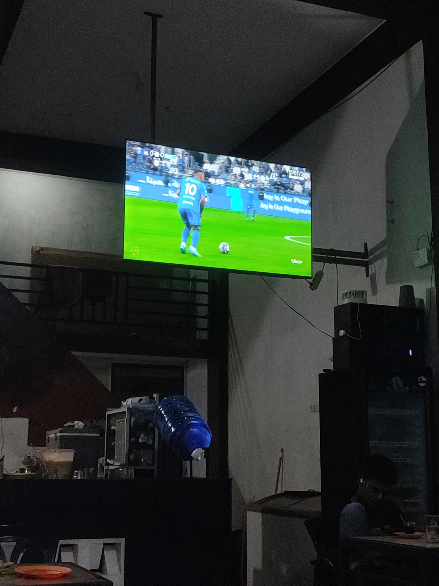 The image shows an indoor scene with a TV displaying a soccer game, featuring a player wearing the number 10. The game is broadcasted by SPOTV2 and Vidio. The environment appears to be a cafe or restaurant setting, with a counter, water dispenser, and a person sitting at a table, possibly in Cotseutui, Indonesia.