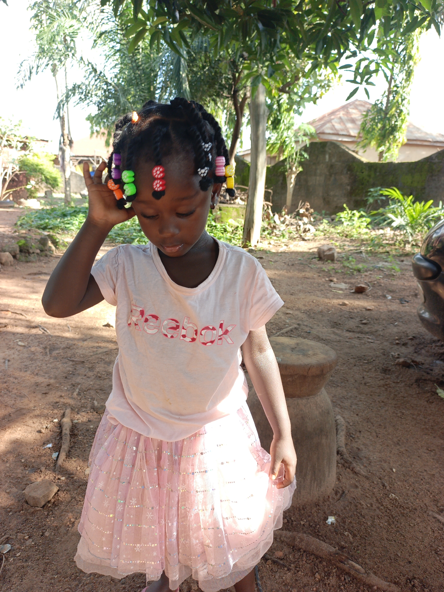An outdoor medium shot captures a young Black girl with braided hair adorned with colorful beads, standing and looking down with her left hand near her head. She wears a light pink t-shirt with a pink Reebok logo and a sparkly pink tutu-style skirt. The background consists of dirt ground, sparse vegetation, trees, and a blurred building with a reddish-brown roof. A dark vehicle is partially visible on the right side of the image. The lighting suggests it is daytime, with sunlight filtering through the trees.