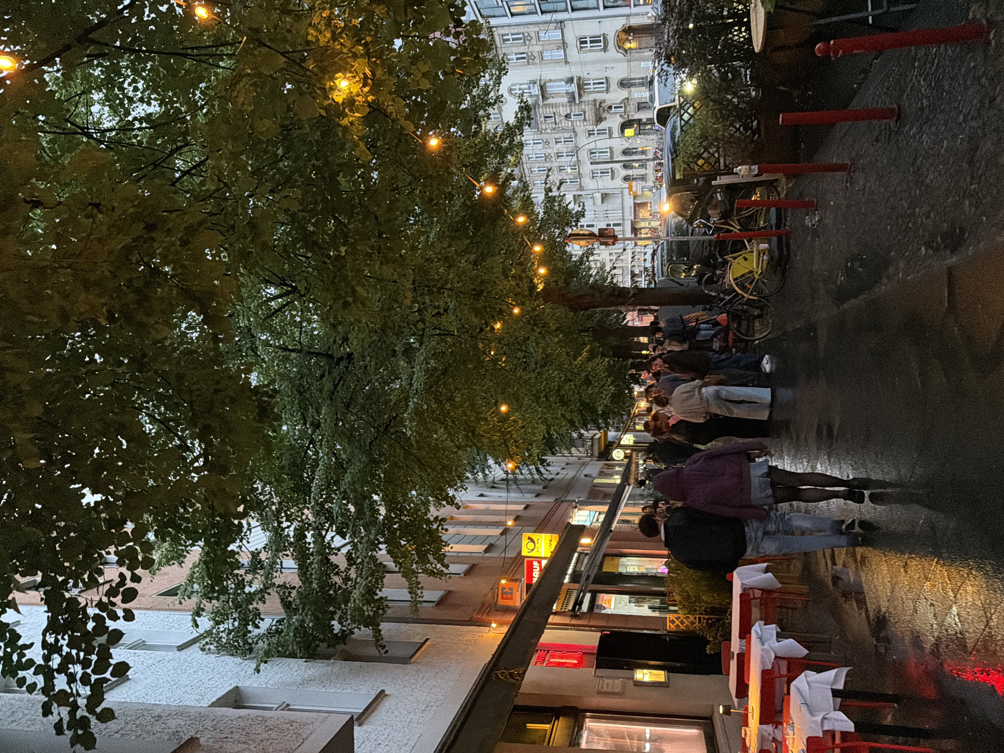 This is a street-level view of a cobblestone street in Berlin, Germany, likely during the evening, given the string lights illuminating the trees and the wet appearance of the pavement, suggesting recent rain or a damp evening. The scene is lined with buildings, some featuring awnings and illuminated signs of businesses, including what appears to be a post office. Trees with dense foliage are planted along the sidewalk, draped with decorative string lights that cast a warm glow. Several people are visible walking along the sidewalk, some in groups and some singly. On the left side of the street, an outdoor seating area with tables and red chairs is set up, some covered with white tablecloths. In the background, cars are parked along the street, and other buildings, typical of Berlin architecture with multiple windows, are visible. The overall atmosphere is that of a lively urban evening scene.