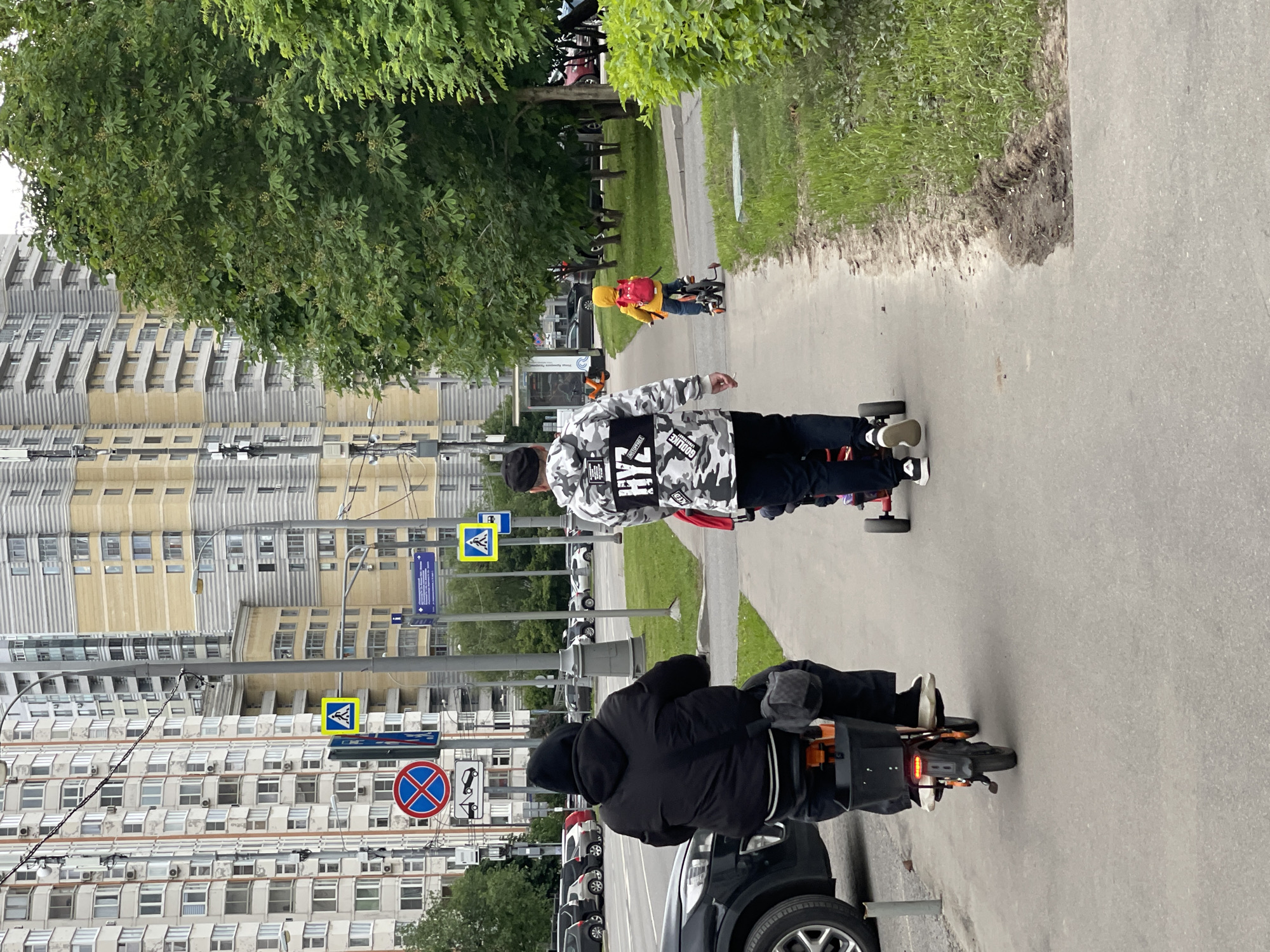 The image depicts a street scene in what appears to be a residential area of Moscow, Russia, as indicated by the architectural style of the apartment buildings in the background. A pedestrian in a white and gray camouflage jacket and black pants is riding a kick scooter along a sidewalk. The person is facing away from the camera, moving in the same direction as a person on an electric bicycle. A third person on a child's bicycle is further down the sidewalk. In the background are apartment buildings, trees, signs and parked cars. A car is parked parallel to the electric bicycle and sidewalk, with its front bumper just behind the electric bicycle. The photo is taken from the side of the street and sidewalk. The light suggests it is daytime.