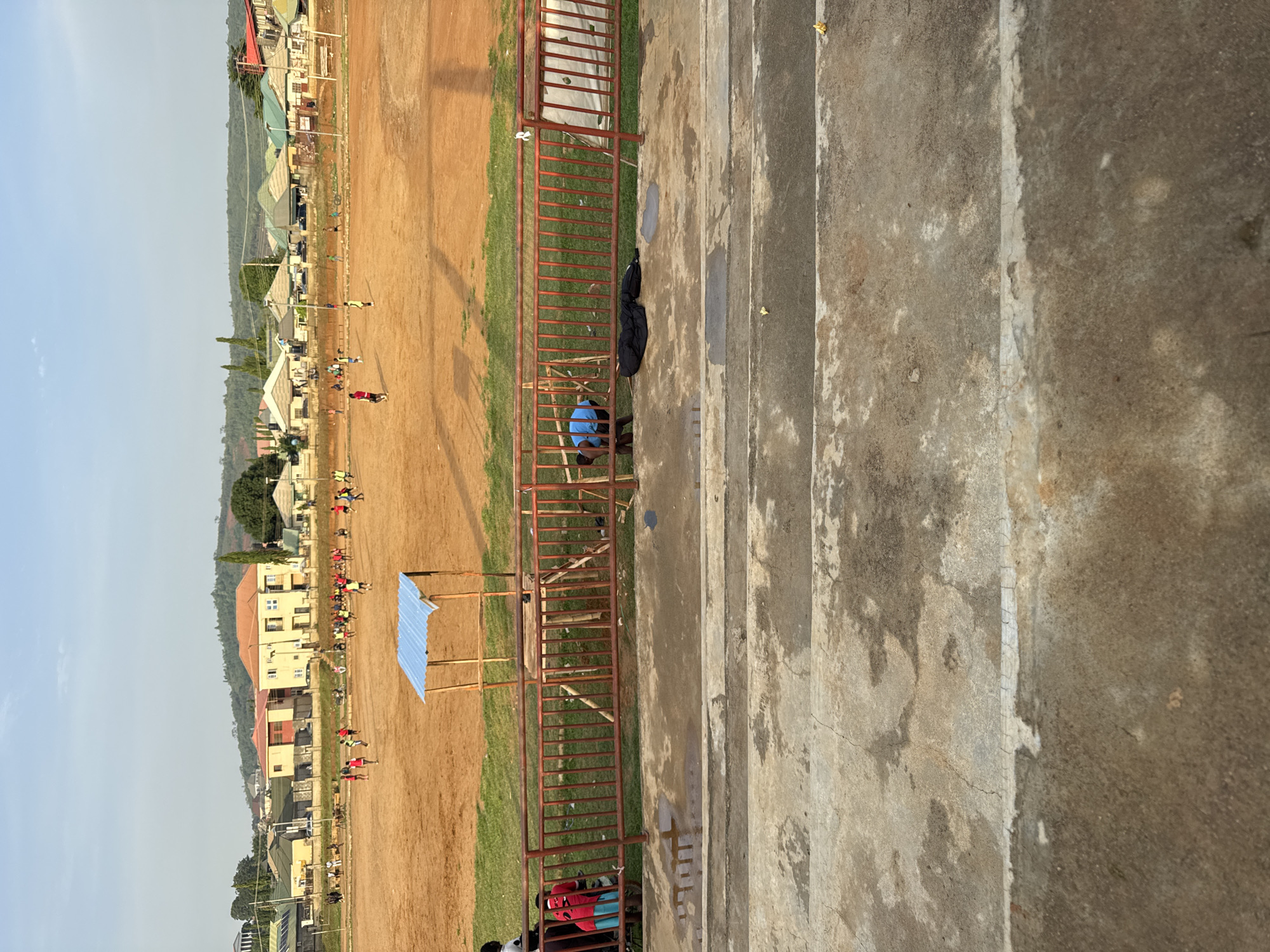 This media file shows an outdoor scene, likely a sports field or track, viewed from an elevated position. The foreground is dominated by concrete steps, showing signs of wear and tear with patches of dirt and discoloration. A dark, folded garment, possibly a jacket or bag, rests on the ground near the edge of the steps.
Beyond the steps, a red metal railing encloses a dusty brown field, which appears to be a dirt track or playing surface. To the left of the field, a thin strip of green grass is visible. Further into the background, a line of buildings with various colored roofs, including red and green, are visible, nestled against a backdrop of lush, green hills. The sky is a pale blue, suggesting it is daytime, likely late afternoon or early morning given the lighting.
Scattered across the field are numerous small figures, some appearing to be children or young adults. They are engaged in various activities, though it's difficult to discern specific actions from this distance and angle. Some appear to be running or walking, possibly participating in a race or training. A small, blue-roofed shelter with a wooden frame is present on the field. The overall impression is a community event or practice taking place in a semi-urban environment in Abuja, Nigeria.