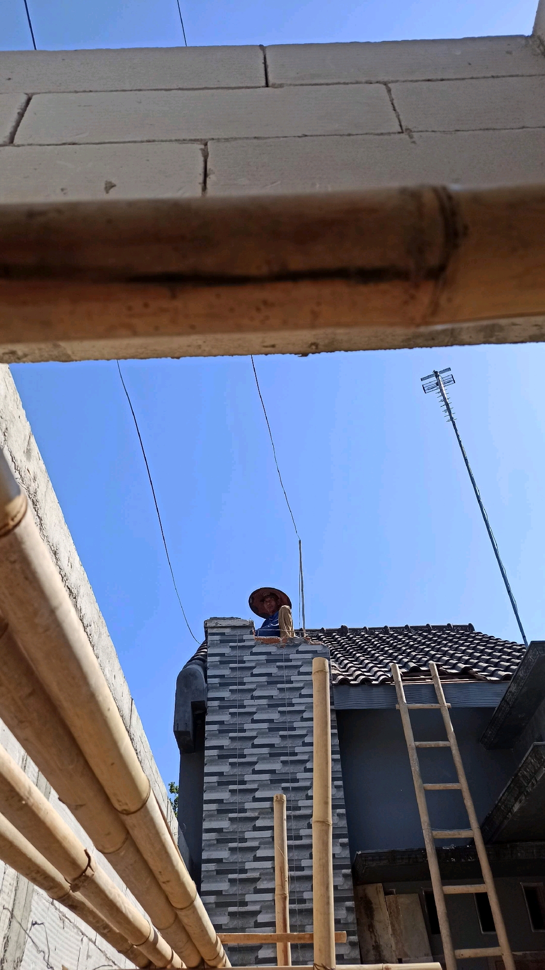 An outdoor, medium-shot image captures a construction scene against a bright blue sky in the city of Ngumpul, Indonesia. In the center, a person wearing a conical hat and a light-colored shirt is perched on a rooftop, appearing to be working on a chimney. The chimney is clad in grey rectangular tiles with a textured pattern. A wooden ladder leans against the side of the building. In the foreground, bamboo poles are arranged horizontally, obscuring part of the view. A tall antenna stands on the right side of the rooftop. The background shows a building with a dark-colored roof and grey walls. The image has a slightly tilted perspective.