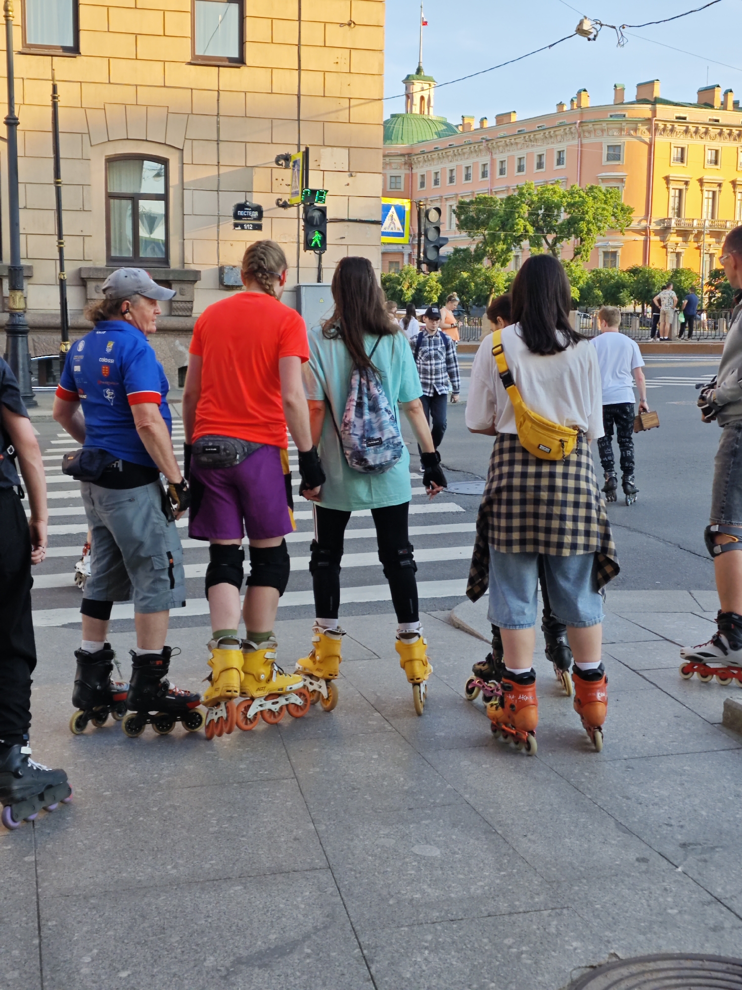 In Saint Petersburg, Russia, a group of people are inline skating on a sidewalk in front of a building. The building is made of light-colored brick and has several windows. A traffic light is visible on the side of the building, showing a green pedestrian signal. In the background, there is a larger, ornate yellow building with green domes, likely a historical landmark. The sky is clear and blue, indicating it is daytime. The people are dressed casually, and some are wearing protective gear like knee pads. The focus is on their inline skates and their movement on the paved surface.