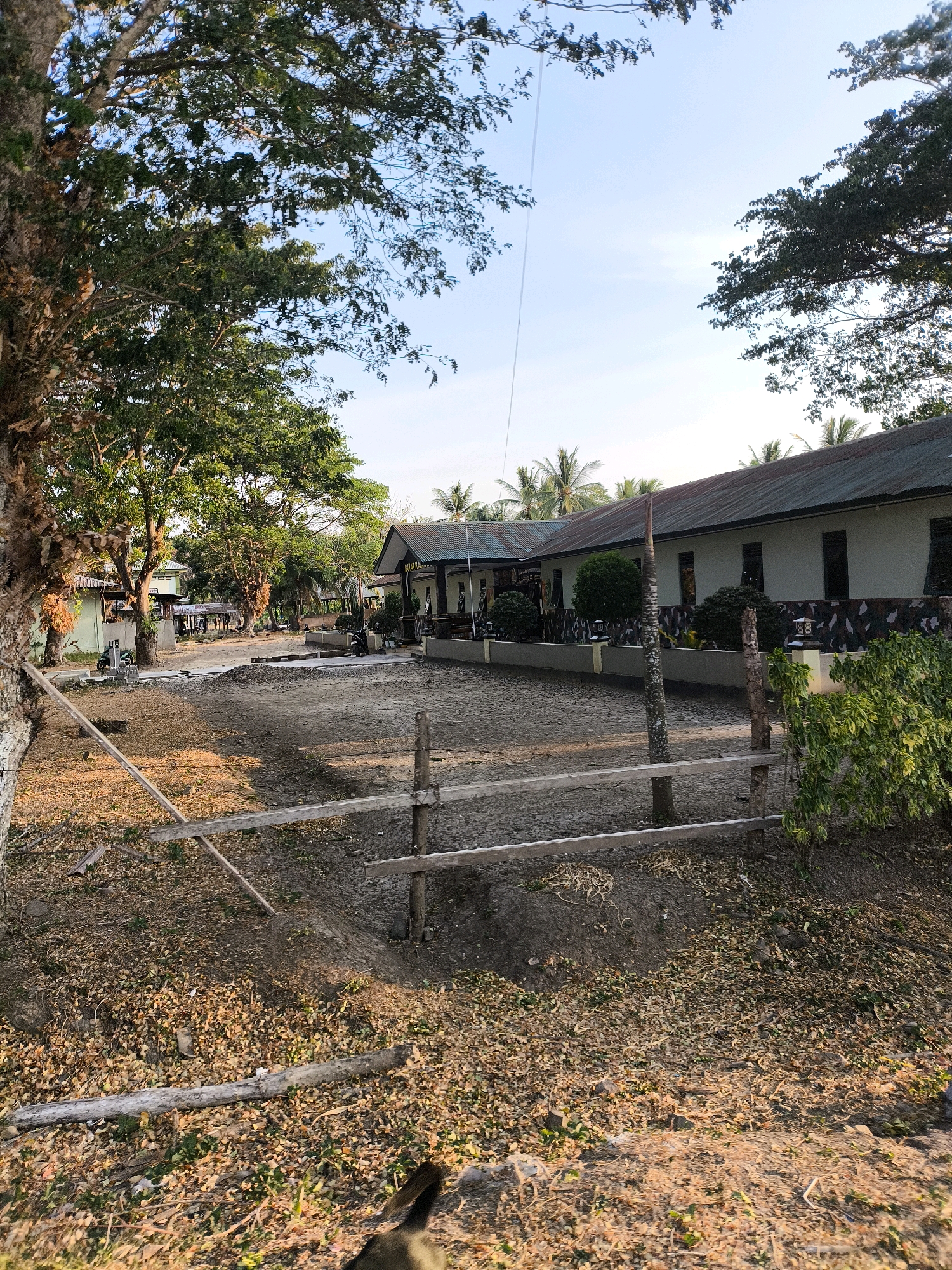 A full, eye-level, outdoor shot depicts a view of a building and its surrounding yard in an Indonesian setting. The scene is captured in the late afternoon, indicated by the warm, golden lighting. The building is long and single-story with a dark, sloped roof and light-colored walls. Camouflage-patterned wainscoting runs along the lower section of the building walls, above a yellow trim. Several windows with dark frames are visible along the facade, and two decorative lamps with pointed tops flank the entrance. A flagpole stands to the left of the building.
In front of the building, the yard is mostly dirt and gravel, with some patches of dry grass and scattered fallen leaves. A low, rough wooden fence made of logs and branches runs horizontally across the foreground, seemingly delimiting an area of disturbed earth and sparse vegetation.
To the left of the building, a large tree with a thick trunk and dense green foliage dominates the frame. The tree's branches extend upwards and outwards, some reaching into the clear blue sky, which is dotted with a few faint clouds. More trees, including palm trees, are visible behind the main building and further into the distance, suggesting a tropical or subtropical environment.
The yard is relatively sparse, with some small bushes and shrubs placed strategically in front of the building and along the fence line. Two motorcycles are parked on the gravel area in front of the building, and another is visible further down to the left. The overall impression is one of a modest, functional building in a tropical climate, possibly a school, administrative office, or residential compound, with a slightly unkempt but lived-in yard.