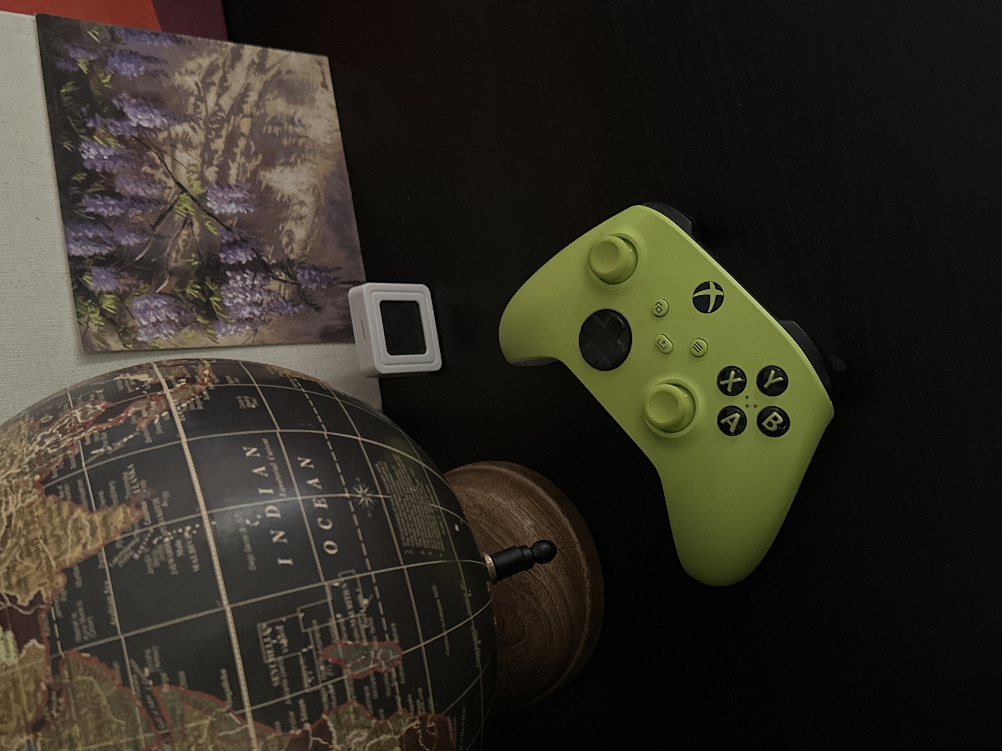 The image shows a still-life composition on a dark, possibly black, surface. The objects include a globe with dark oceans and continents in muted colors, a small white square device with a black screen, a painting of purple flowers on a canvas, and a lime green Xbox controller. The globe is positioned in the lower left of the frame. The controller is placed in the upper right portion of the image. The other two items are more to the left, closer to the globe. The image appears to be shot from a high-angle perspective with no one in view. The location in Šušanj, Montenegro is unknown from the image alone.
