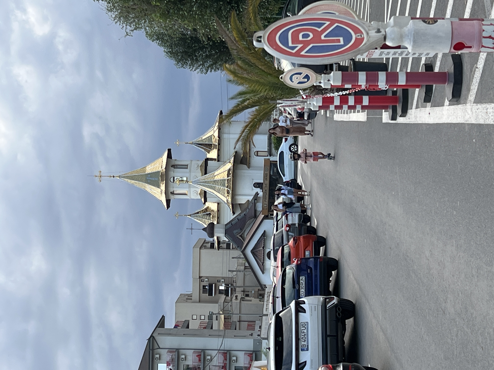 This image depicts a street scene in Năvodari, Romania, with a prominent Orthodox church featuring golden domes in the background. A row of parked cars lines the right side of the asphalt road. In the foreground, a child rides a scooter on the sidewalk, accompanied by two women walking behind. Several "no parking" signs are visible along the sidewalk, along with red and white bollards. Buildings, likely apartment blocks, are situated to the left of the church. The sky is overcast with dramatic clouds.
