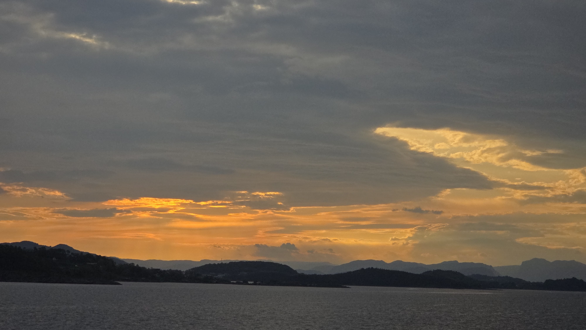 The image presents a dramatic sunset over a tranquil body of water, likely a fjord or sea, set against a backdrop of distant hills and mountains in Randaberg, Norway.
The sky dominates the upper two-thirds of the frame, showcasing a striking contrast between dark, heavy grey clouds and brilliant sunset colors. Thick, textured clouds stretch across the upper sky, appearing dark and somber. Below these, the setting sun illuminates the clouds from behind and underneath, creating intense bands of fiery orange, golden yellow, and warm red light. Several breaks in the cloud cover allow bright, almost luminous yellow light to burst through, especially prominent in the lower left and upper right portions of the sky, where the light seems to glow from within the clouds.
Along the horizon, a series of dark, silhouetted mountains and hills delineate the landmass. These landforms appear as deep blue-grey and black shapes against the vibrant sky, exhibiting varied contours and elevations that suggest a rugged coastal or fjord landscape. The lower quarter of the image is filled with the calm, dark surface of the water, which reflects the muted light from the sky above. The water shows subtle textural variations, hinting at gentle movement.
The time of day is unmistakably sunset, characterized by the rich, warm color palette. The weather appears cloudy, yet the dramatic illumination of the clouds creates a powerful and majestic scene. The overall atmosphere is one of serene natural beauty and dramatic light. There are no people, man-made objects, or visible text in the image.