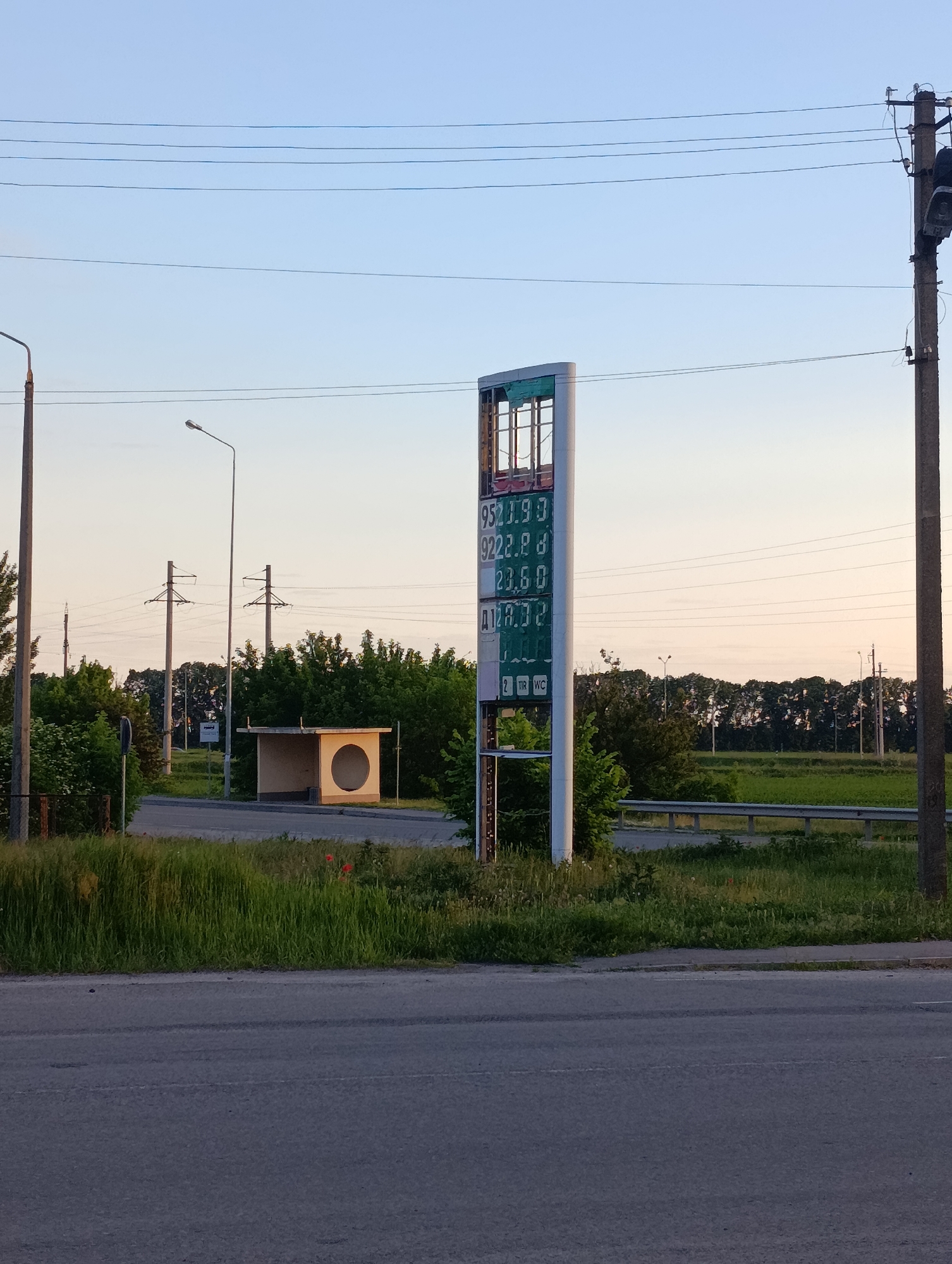 This media file captures a quiet roadside scene in Krasnohorivka, Ukraine, bathed in the soft light of either dawn or dusk. The sky transitions from a pale, warm orange-pink at the horizon to a clear, light blue overhead, indicating fair weather.
In the foreground, an asphalt road runs horizontally, leading to a grassy verge where a few small red flowers bloom. The midground is characterized by several infrastructure elements. To the left, a concrete bus stop or shelter with a distinctive circular cutout on its front face stands beside a tall streetlight. Behind these, utility poles support multiple power lines that stretch across the frame.
A prominent feature is a tall, narrow sign, likely from a gas station, positioned centrally. Its upper portion appears somewhat damaged. The digital displays on the sign show what are presumably fuel prices:
- "9521.99" (likely for 95 octane fuel)
- "9222.88" (likely for 92 octane fuel)
- "23.50"
- "Д 127.32" (with "Д" commonly representing Diesel in Cyrillic-speaking regions).
Below the prices, smaller icons indicate available services: "2", "TIR", and "WC" (toilet).
Behind these structures, dense green foliage, including bushes and trees, forms a natural barrier along a slightly elevated section of land, where a metal guardrail is also visible. The background reveals a flat, open field stretching towards a distant line of trees under the fading sky. There are no people visible, contributing to a sense of calm and stillness in the scene. The setting suggests the outskirts of a town or a rural road.