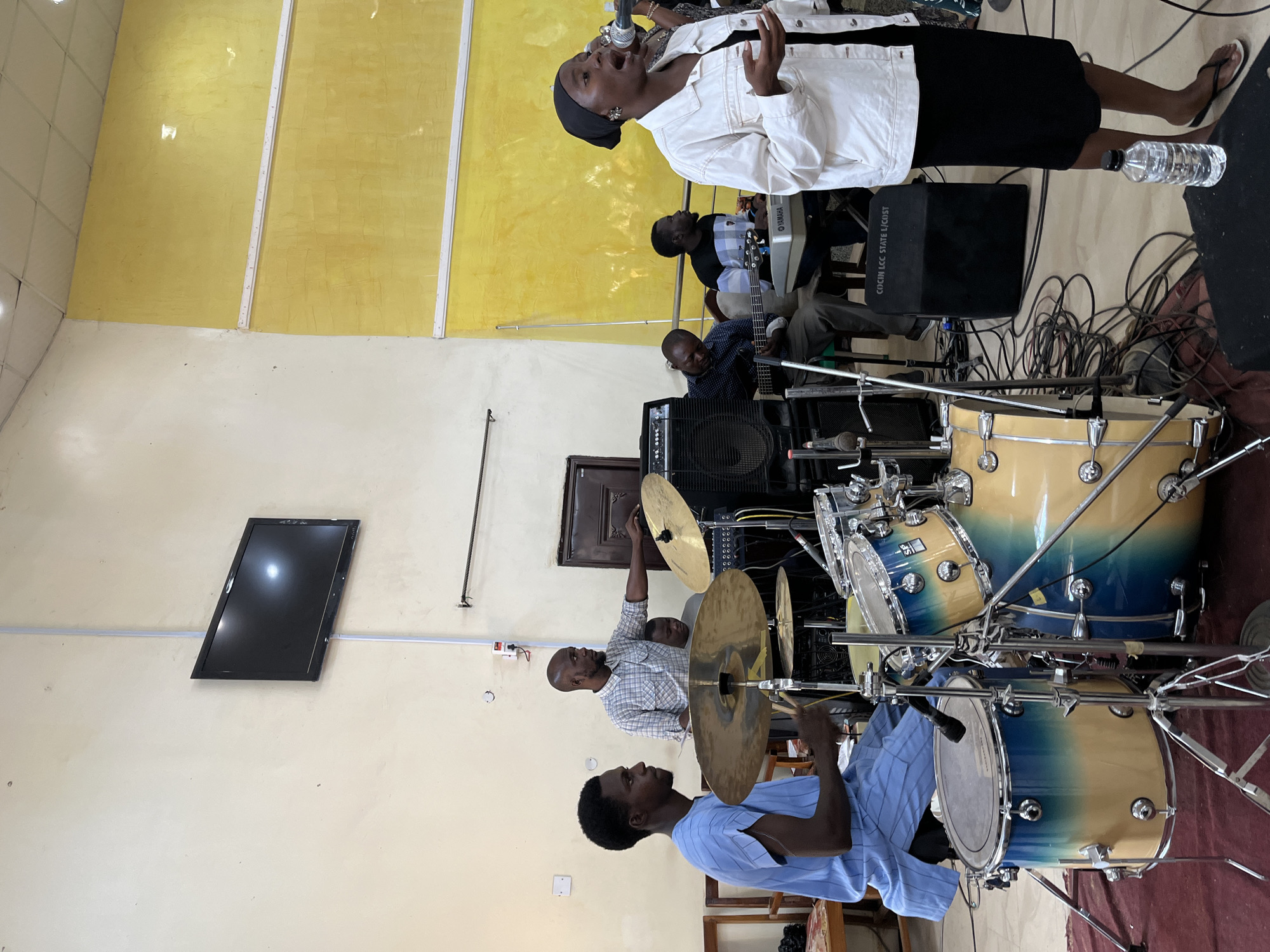 The image captures a live musical performance likely taking place in a church or religious setting in Jos, Nigeria. A female singer dressed in a white jacket, black skirt, and a black head covering stands in front of a microphone, energetically singing. Behind her are musicians playing instruments. There's a bass guitarist, a keyboardist, and two drummers playing a full drum kit. A sound amplifier with "COCHIN LCC STATE L/CRIST" printed on it is visible near the keyboardist. A television screen is mounted on the wall, and the room has yellow paint. The scene is filled with musical equipment, including amplifiers, cables, and the drum kit itself. There's also a water bottle sitting on the floor.