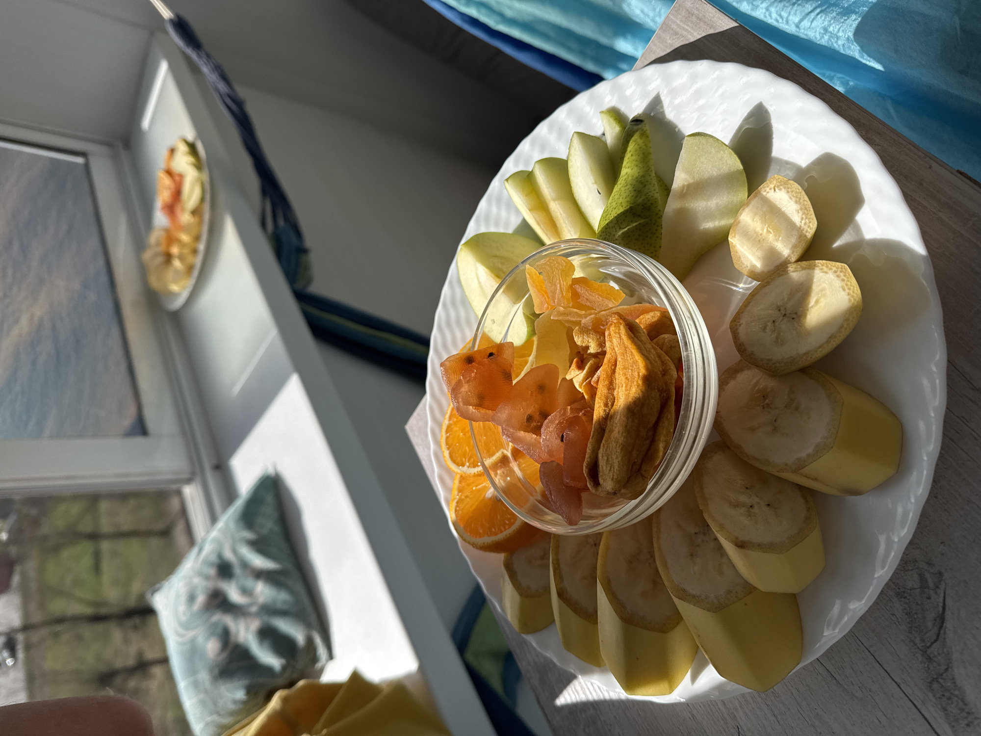 A fruit platter is centrally featured, showcasing an assortment of fresh and dried fruits. A large, white, scalloped-edge plate holds slices of green and yellow apples, green pears, and bananas arranged around a smaller, clear glass bowl. Inside the glass bowl are various dried fruits, including orange-colored dried apricots and other golden-brown, elongated dried pieces. Surrounding the bowl are also slices of tangerine or orange.
The platter rests on a light-colored wooden surface, possibly a table or windowsill. To the left, a window provides bright, natural light, creating distinct, long shadows across the plate and the surface. Through the window, a blurred image of another plate with fruit is faintly visible. On the right side of the image, vibrant blue curtains add a splash of color to the background. A patterned blue and white cushion is partially visible below the window on the left.
The strong, direct sunlight suggests it is daytime, likely morning or late afternoon, given the sharp shadows. The scene depicts a prepared snack or light meal in a bright, indoor setting, possibly a home. There is no visible text in the image.