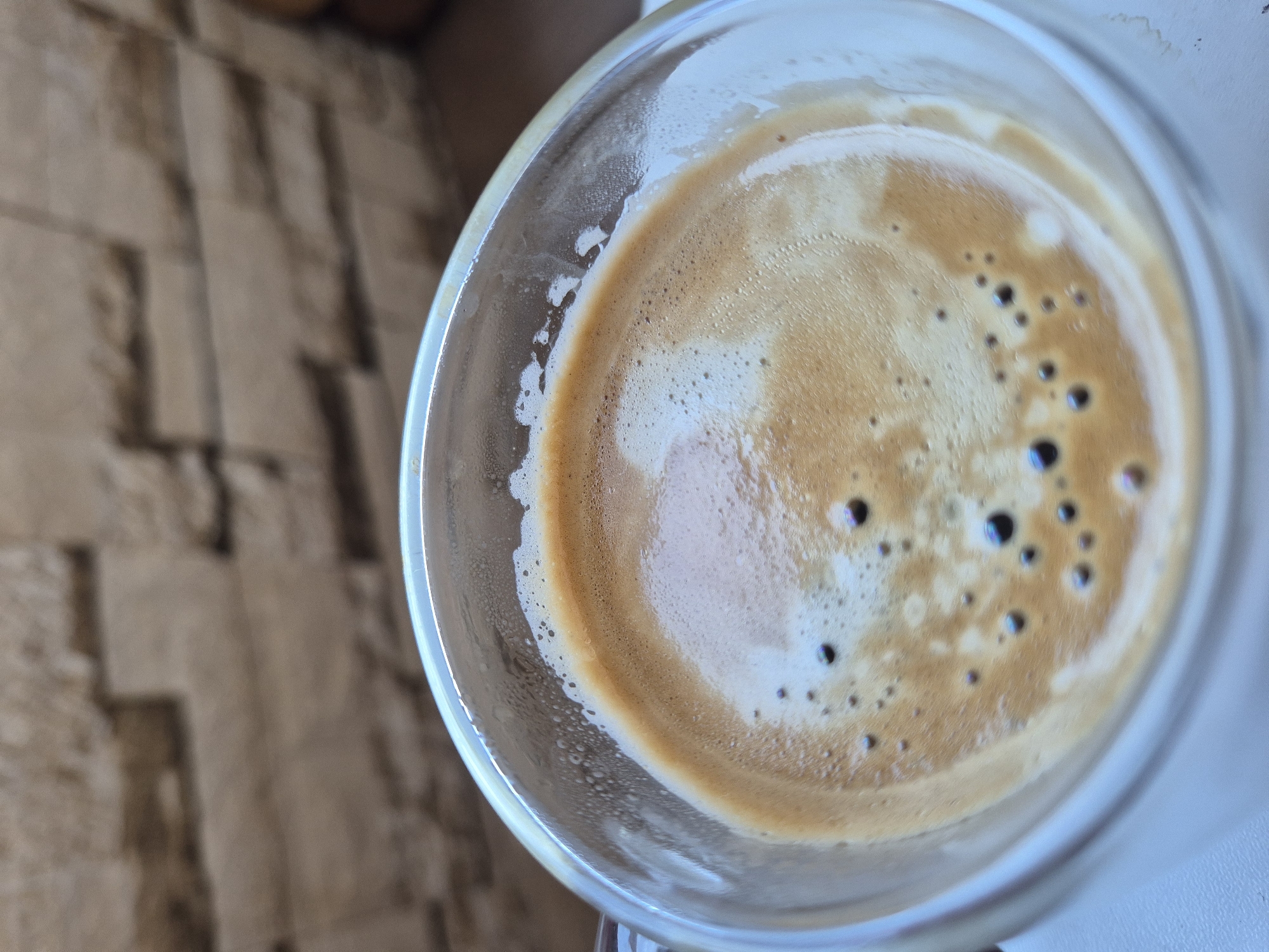 This media file shows a close-up, top-down view of a glass of espresso. The espresso has a rich, creamy crema on top, with small bubbles visible. The background is blurred and appears to be a textured surface, possibly a wall or a table, with a neutral brown color. The lighting suggests it might be daytime, as there is a bright light source creating reflections on the glass. There are no people or any specific location cues visible in the image. The focus is entirely on the coffee, highlighting its texture and color. The overall impression is one of a simple, still moment, possibly capturing the preparation or enjoyment of a coffee drink.