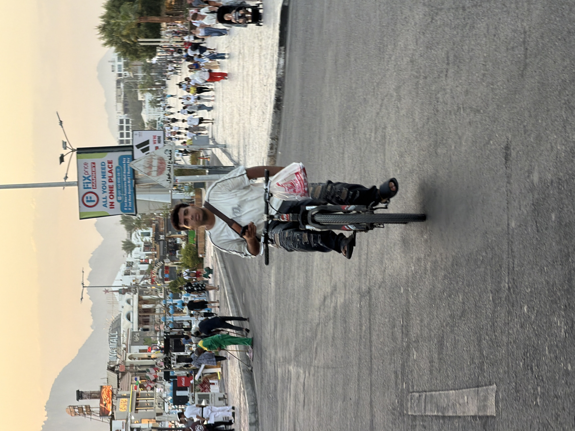 The image shows a street scene in Sharm El Sheikh, Egypt, likely in the Naama Bay area, given the presence of signs for "Mina Mall" and "Fix Price Market" with addresses indicating Naama Bay. The foreground is dominated by a man riding a bicycle with a plastic bag hanging from the handlebars. He is wearing a white t-shirt, distressed jeans, and dark sandals. The man is looking forward as he cycles on an asphalt road.
In the background, a pedestrian walkway is visible, filled with people walking in various directions. Some individuals are dressed casually, while others appear to be tourists. Among the crowd, a person is pushing a stroller with a baby. To the left of the pedestrian walkway, shops and buildings with signage are lined up, suggesting a commercial and tourist hub. The buildings have an architectural style typical of the region, with some featuring balconies and awnings. Palm trees are interspersed along the walkway and behind the buildings, indicating a warm climate.
The sky in the background is a soft orange and yellow, suggesting either sunrise or sunset. Mountains are visible in the distance, providing a scenic backdrop to the urban environment. The overall scene depicts a lively street scene in a popular tourist destination in Egypt, with daily activities like cycling and shopping taking place.