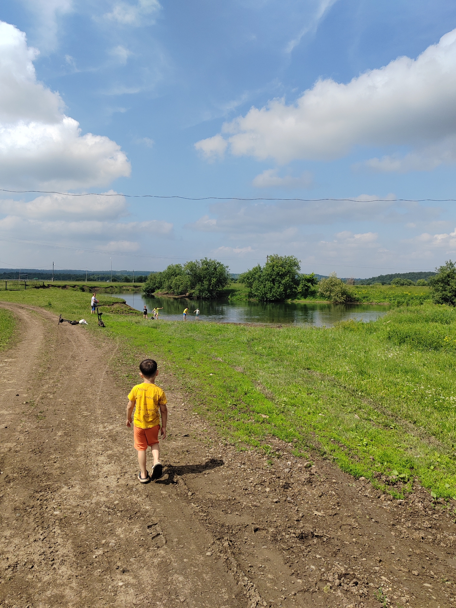A young boy in a yellow t-shirt and orange shorts walks away from the camera on a dirt path. In the background, several other children and an adult are near a body of water, with trees and a grassy field surrounding it. Two scooters and a white object are lying on the grass near the path, to the left of the frame. The scene appears to be a park or natural area on a sunny day, with scattered clouds in the blue sky.
