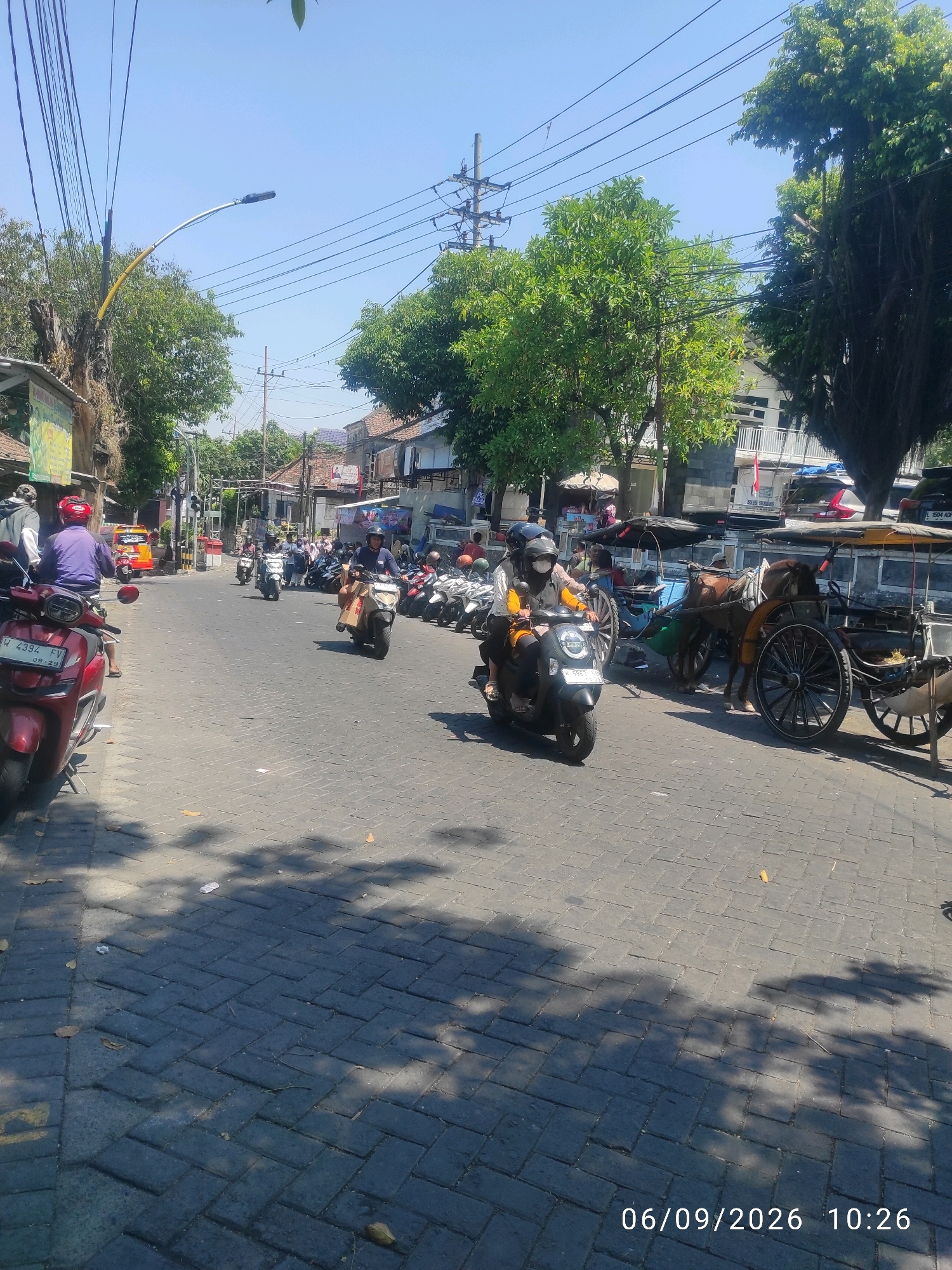 The image shows a street scene in Kebomas, Indonesia, on a sunny day. The street is paved with interlocking bricks and lined with buildings and trees. Several people are on motorcycles, and there are also horse-drawn carriages visible. The overall atmosphere is one of a bustling urban environment.
In the foreground, a red motorcycle is parked on the left side of the street. Behind it, a person wearing a purple shirt and a red helmet is seated on another motorcycle.
Further down the street, a person wearing a yellow shirt and a helmet is riding a black scooter. Another person, also wearing a helmet, is seated behind them.
On the right side of the street, a horse-drawn carriage is parked. The carriage is brown and has large, spoked wheels. It is decorated with some yellow trim and appears to be carrying passengers.
Several other motorcycles are parked along the side of the road, and more people are seen on motorcycles in the background. Buildings line both sides of the street, with some appearing to be shops or residences. Trees are interspersed throughout the scene, providing shade. Power lines are visible overhead.
The image is dated "06/09/2026 10:26", suggesting it was taken on September 6, 2026, at 10:26 AM.