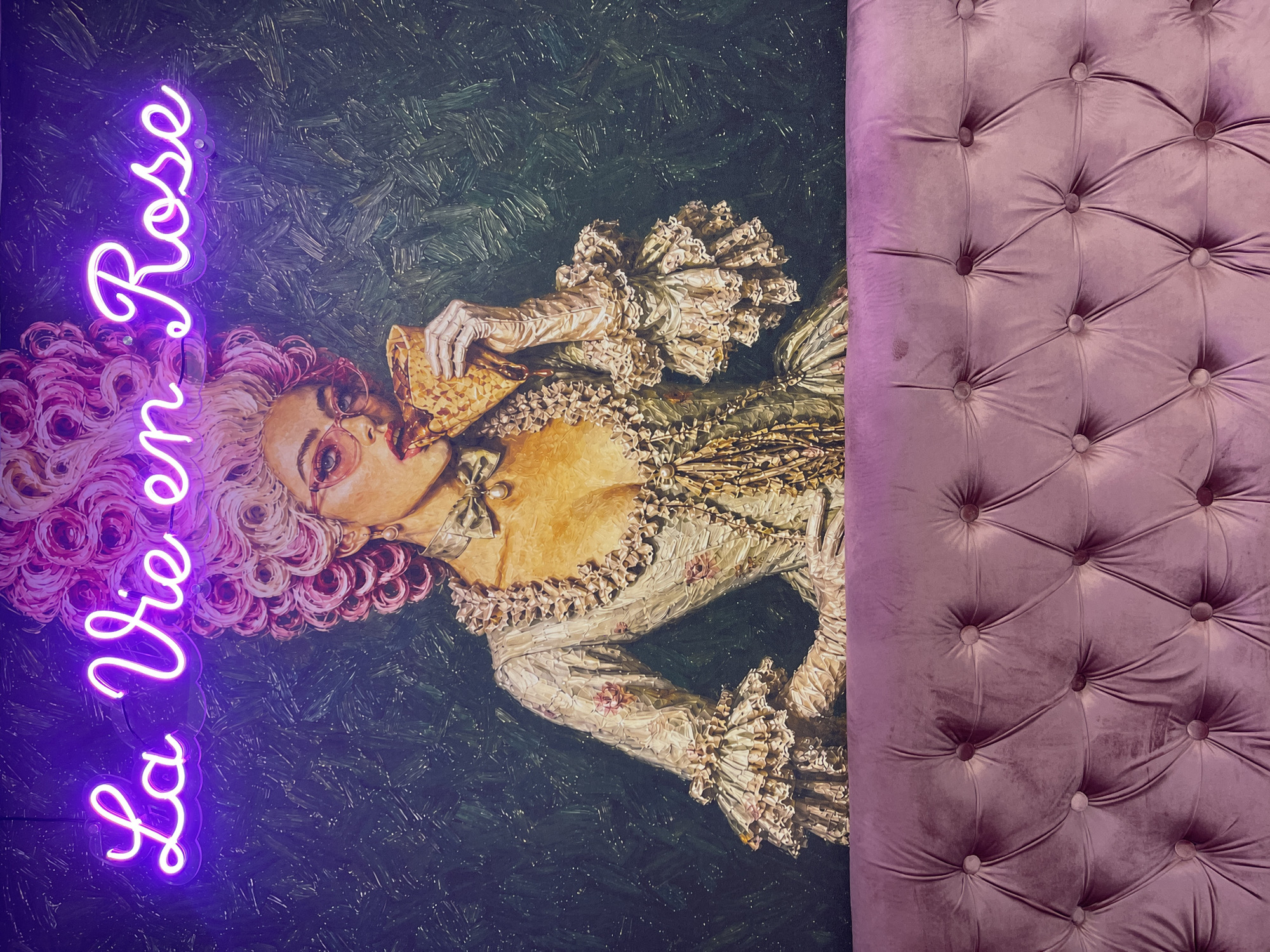 The image shows a wall adorned with a neon sign that reads "La Vie en Rose" in purple script. Below the sign is a large painting of a woman in a lavish historical dress and a towering pink wig, wearing pink-rimmed glasses and eating a waffle cone. The background of the painting is a dark, textured pattern. In the foreground, a tufted purple velvet sofa is visible. The scene appears to be indoors, likely a restaurant or cafe. There is no indication of the location being in North Kuta, Indonesia.