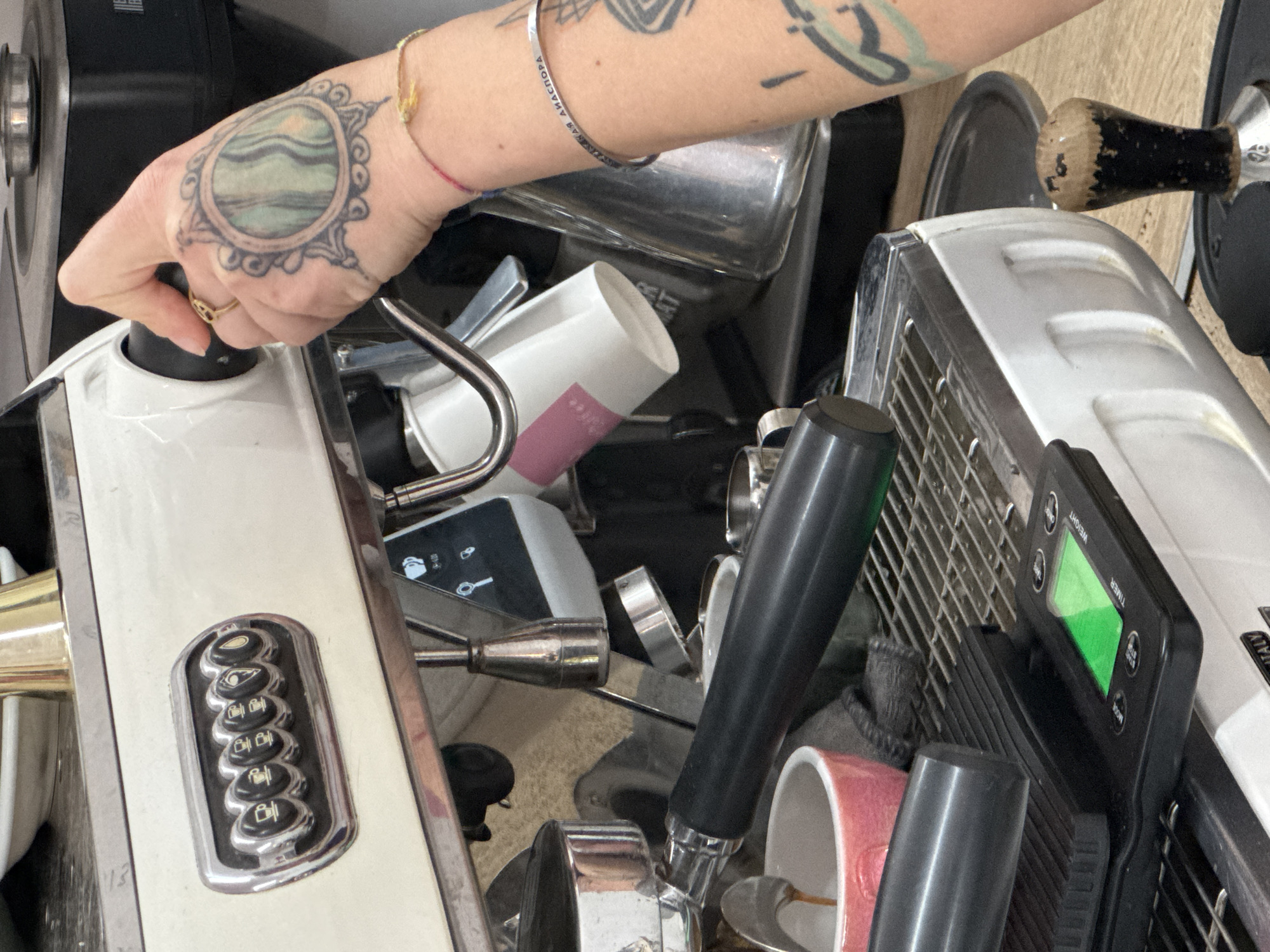 A close-up shot shows a person operating a professional espresso machine, likely in a cafe setting in Bar, Montenegro. The person's left hand, adorned with a ring and a circular tattoo, is turning a knob on the machine. Their forearm displays additional tattoos and bracelets. The machine is a white, stainless steel model with chrome details, featuring a row of buttons and a digital display. Various coffee-making accessories, including a portafilter, tamper, and mugs, are crowded around the machine. A disposable coffee cup is also visible. The overall setting suggests a busy coffee preparation area.