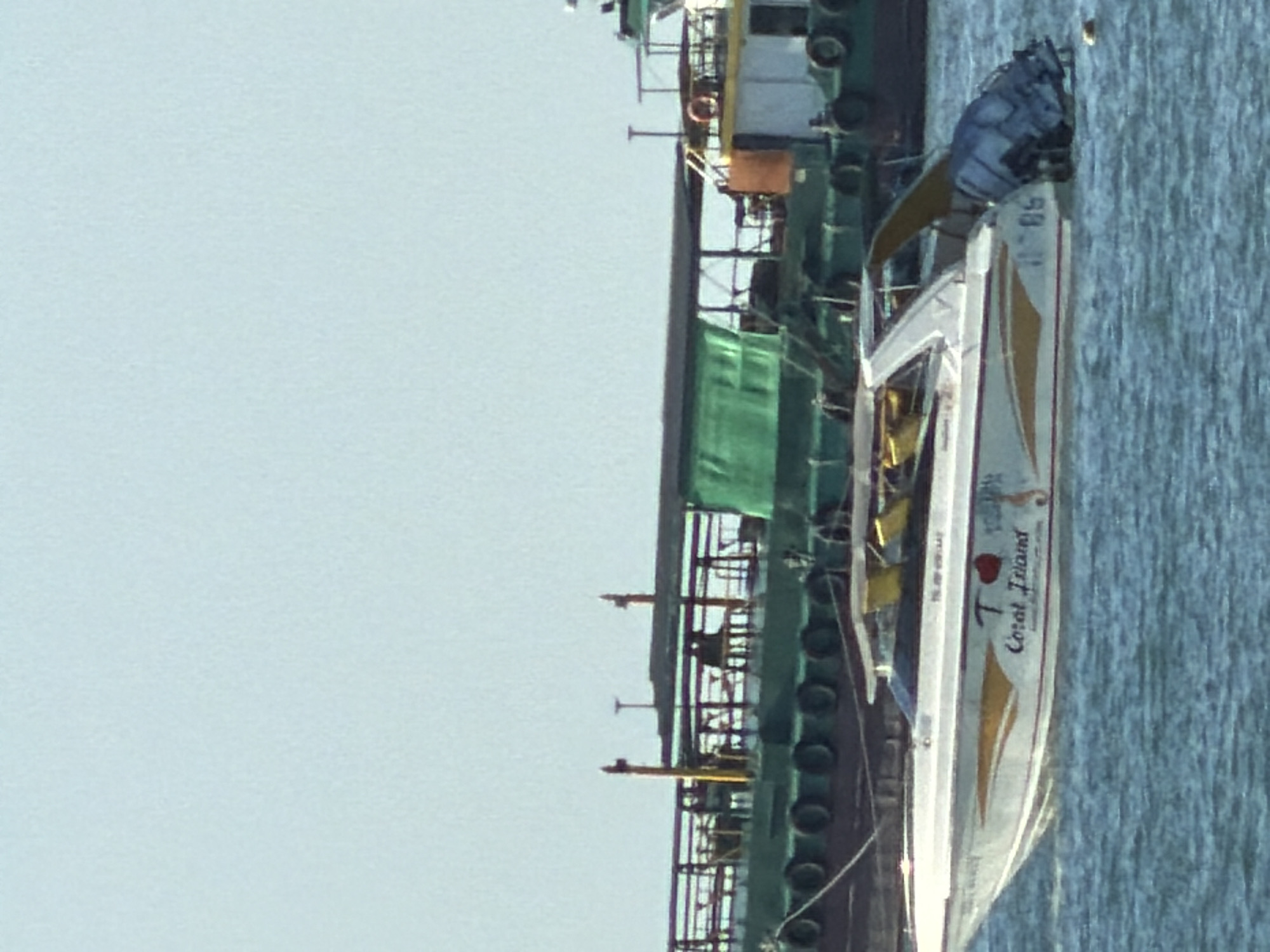In Pattaya City, Thailand, a white speedboat with yellow and red trim is moored in the water. The boat is named "Coral Island" and has the registration number "JB 85" visible on its stern. The speedboat is positioned in the foreground, while a large green cargo ship with a white superstructure is in the background. The water is a deep blue, and the sky is a pale blue with a few wispy clouds. The scene appears to be a calm day at sea.