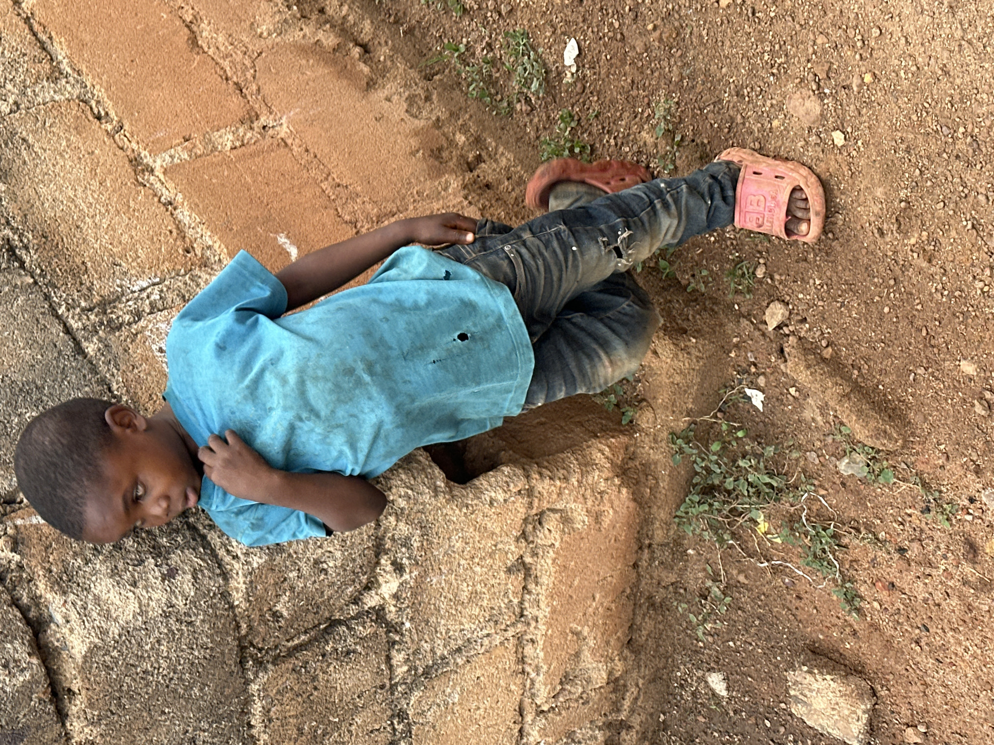 A young boy, estimated to be between 4 and 6 years old, stands leaning against a rough, textured wall made of weathered bricks and large stones. He is wearing a faded turquoise t-shirt with several visible holes and a pair of distressed denim jeans. On his feet, he has bright pink slide sandals. The ground around him is composed of reddish-brown dirt with small pebbles and sparse green vegetation. The scene appears to be outdoors, likely in a semi-urban or rural setting within Gura Top, Nigeria, based on the architectural style and the natural surroundings. The boy is looking to his left, with his left hand on his shirt.