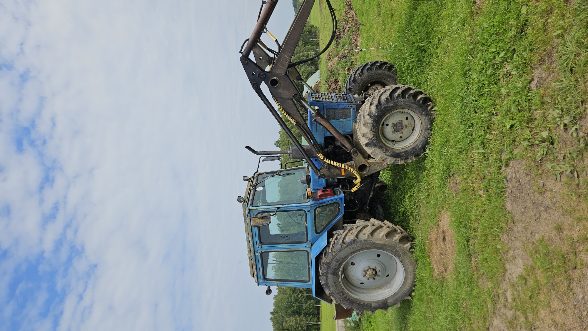 A blue tractor with a front loader attachment is parked on a grassy field. The tractor is facing to the right, with its cabin visible. The sky above is partly cloudy. In the background, there are trees. The scene appears to be in a rural area, possibly in Kapūņi, Latvia, given the context provided. There are no people visible in the image.