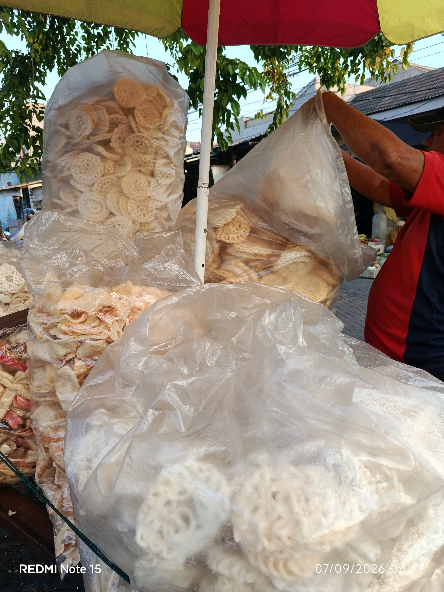 The image depicts an outdoor market stall in Keboansikep, Indonesia, likely a street-side vendor. The foreground is dominated by large, transparent plastic bags filled with various types of white and off-white crackers. One bag, to the left, contains circular crackers with intricate, lace-like patterns. Another bag, centrally located, holds flatter, irregularly shaped crackers that appear golden brown and crispy. A third, larger bag in the foreground also contains white, circular crackers, some appearing somewhat powdery. In the background, a person wearing a red and navy blue shirt is seen reaching into a large plastic bag, seemingly adjusting or inspecting its contents. The stall is partially covered by an awning made of red, yellow, and green fabric. Behind the stall, a building with a tiled roof and some greenery are visible, suggesting a residential or semi-urban environment. The overall scene suggests a vendor preparing or displaying snacks for sale.