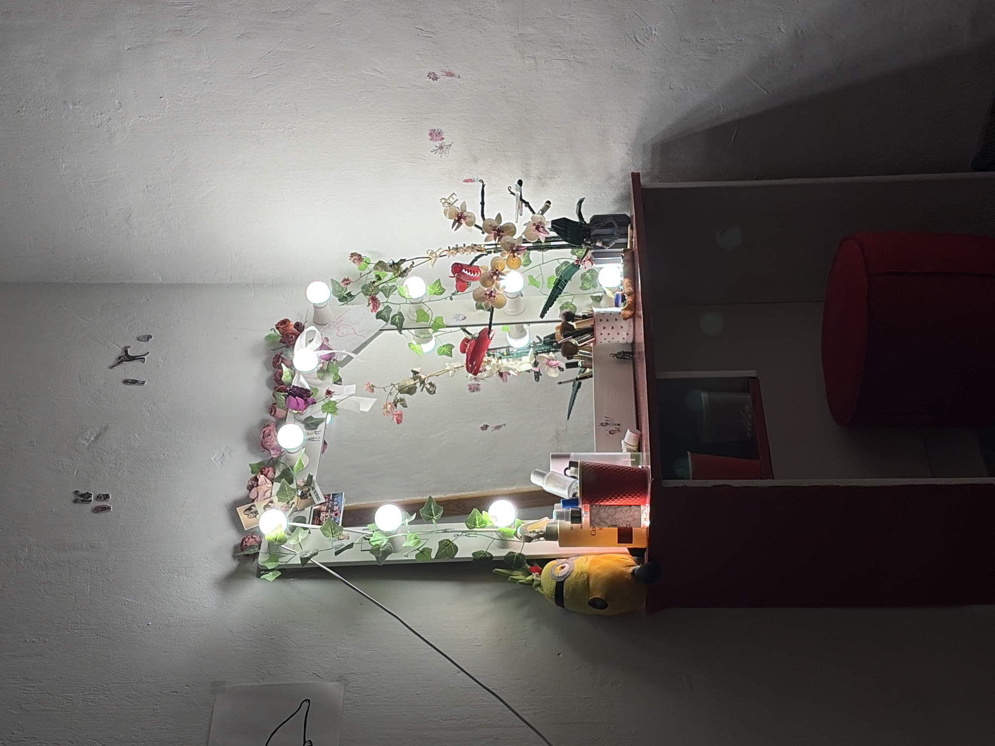 The image depicts an interior wall, likely within a dwelling, decorated with a mirror surrounded by floral and light embellishments. The mirror reflects the room, adding depth to the scene. A strand of lights, interspersed with artificial foliage and flowers, encircles the mirror's perimeter. A Minion plush toy peeks from behind the mirror’s shelf, which holds makeup and other cosmetic items. The adjacent shelving unit is composed of horizontal planes of furniture and red objects.