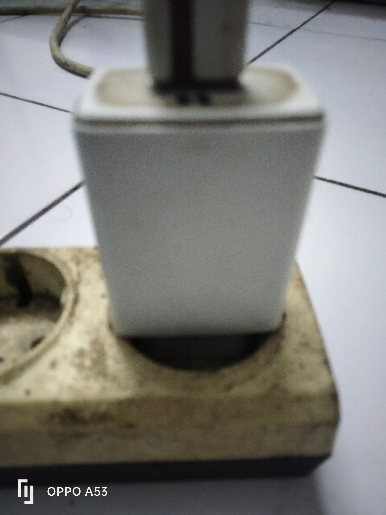 A white power adapter is plugged into a beige, dual-socket electrical outlet, which is situated on a white tiled surface. A grey cable extends from the top of the power adapter. The outlet appears worn and dirty, with dark marks and dust accumulated on its surface. The power adapter is rectangular and white, with a darker rectangular element visible at its top. The scene is dimly lit, and the image is slightly out of focus, particularly the power adapter. The bottom left corner of the image displays a logo that reads "OPPO A53". The scene depicts a common household electrical setup.