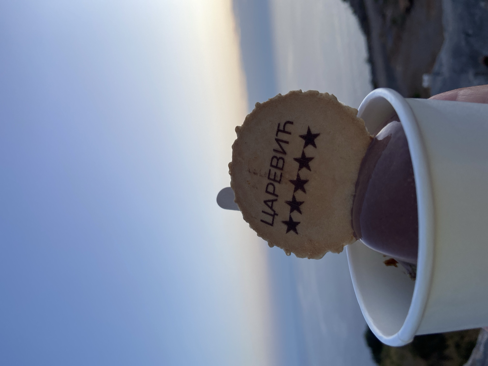 In the city of Kovačko Polje, Montenegro, a close-up shot captures a white paper cup filled with chocolate ice cream. A round biscuit, emblazoned with the Cyrillic word "ЦАРЕВИЋ" and four black stars, is perched atop the ice cream. The background reveals a hazy expanse of the sea meeting the sky, with a faint silhouette of a mountainous landscape on the right. The lighting suggests it is either dawn or dusk.